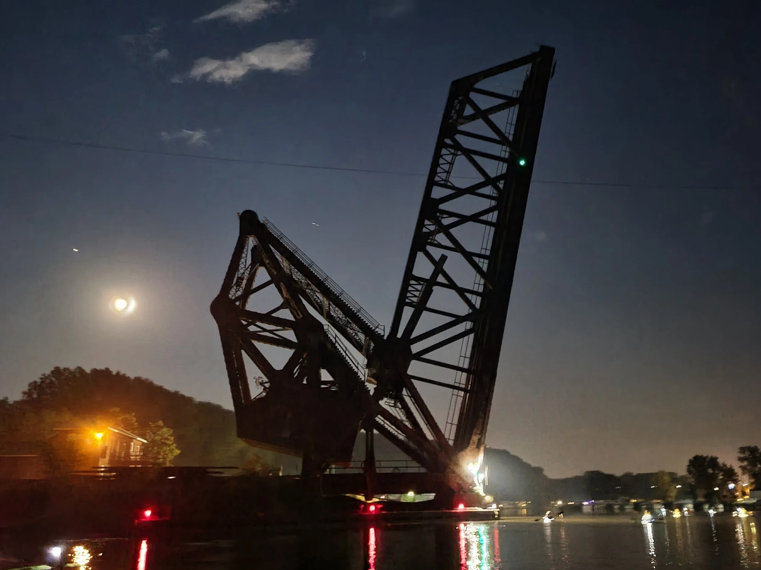 Nighttime image of a large, tall crane structure with a green light, silhouetted against the moon and a dark sky with clouds. Trees and streetlights are visible below, with reflections on the water.