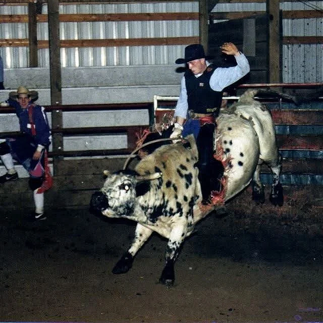 A person dressed in cowboy attire riding a fake or deceased animal prop, which resembles a large animal with spots, inside a barn or arena. A child is seated on the left side.