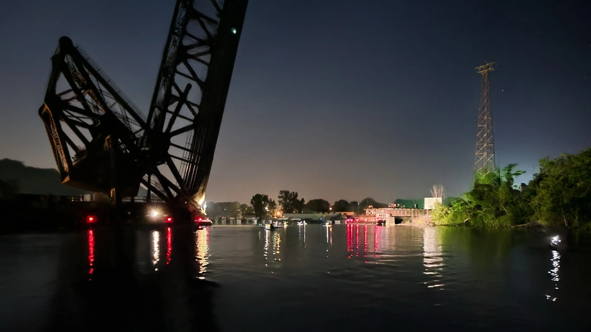 Nighttime scene of a canal with an overturned crane on the left and a tall radio tower on the right, with trees, water reflections, and some lights in the distance.