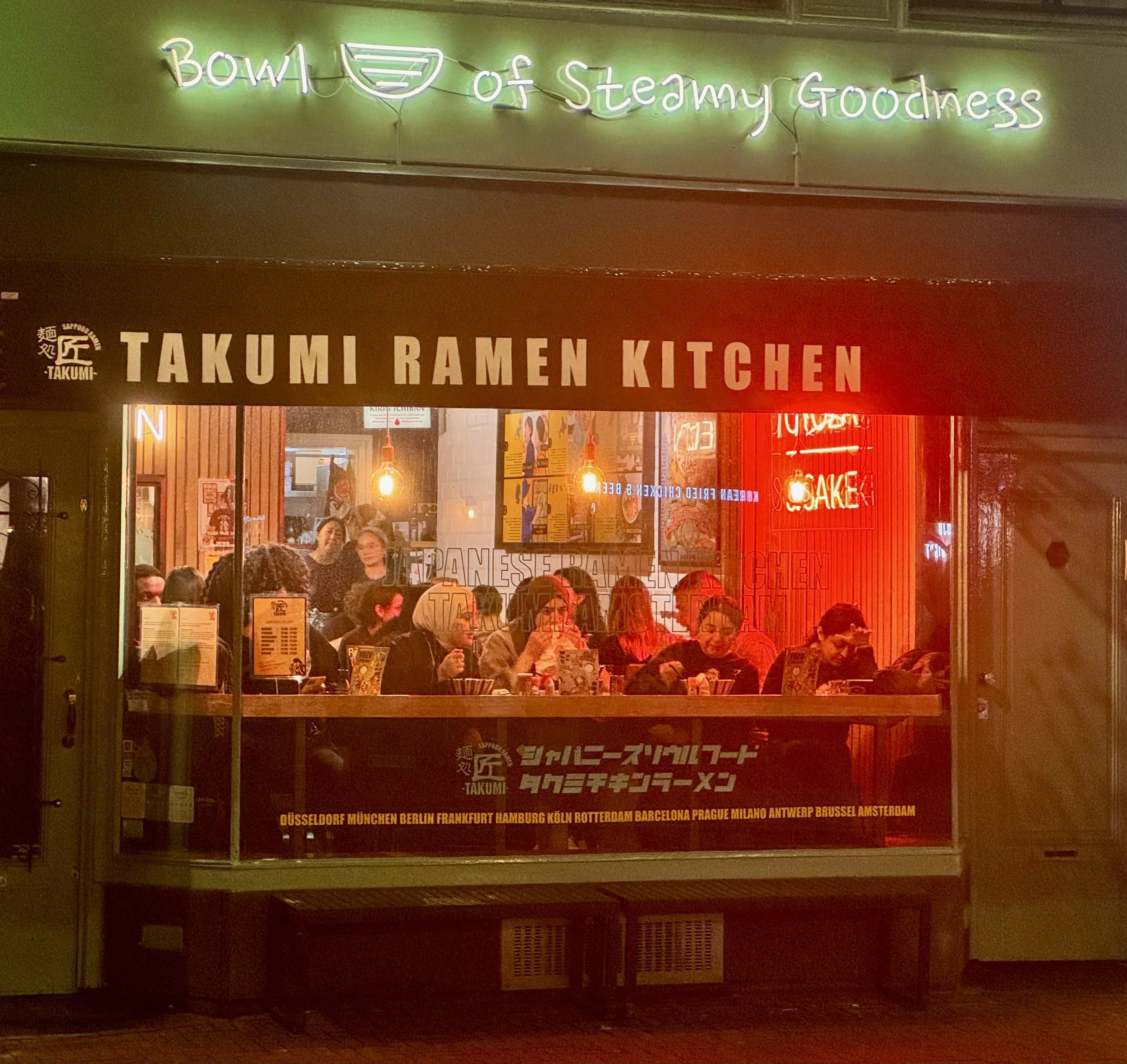 Neon-lit ramen restaurant storefront bustling with diners at night