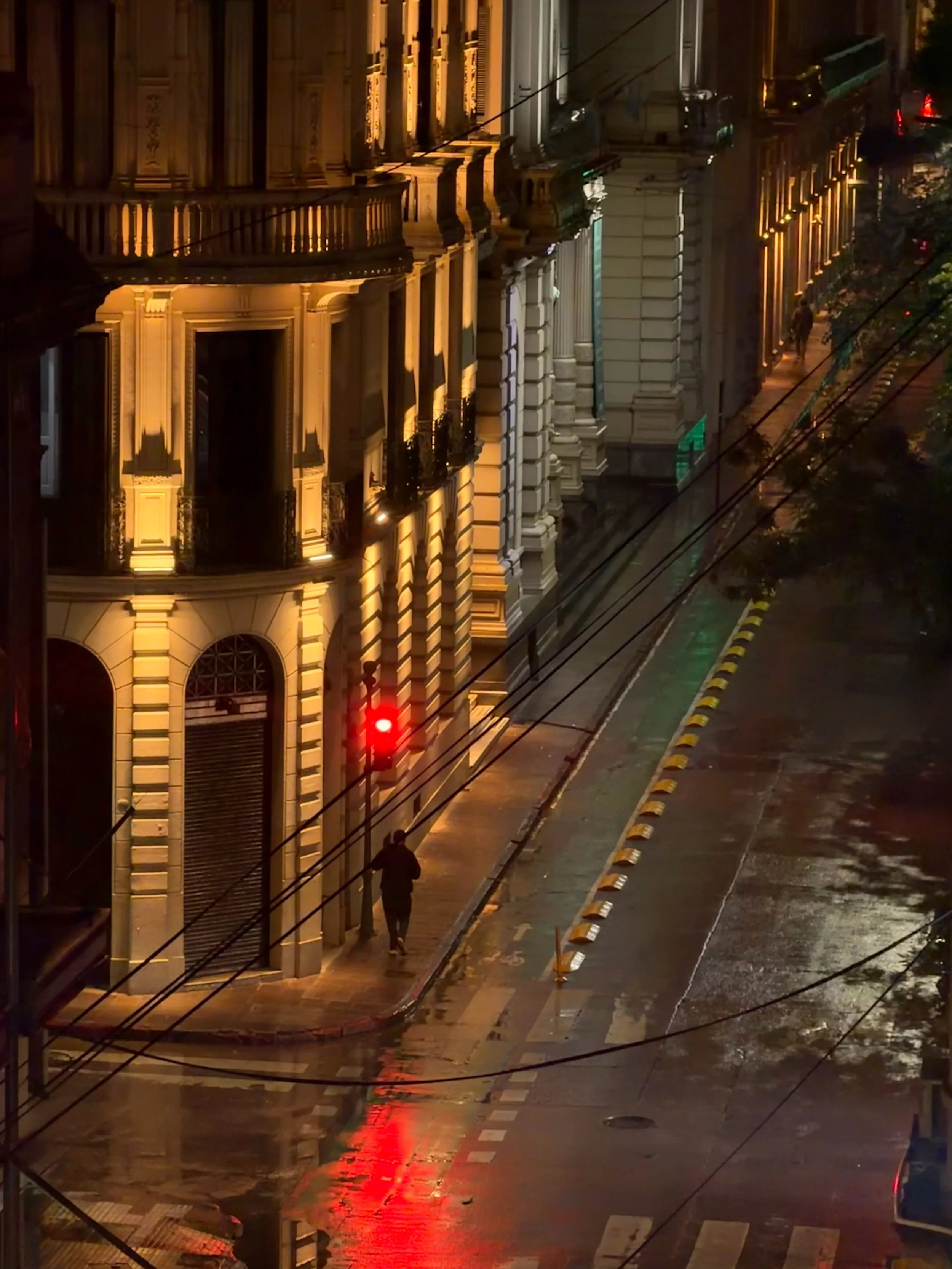 Lone figure walking a rain-slicked city street lit by warm lights and a red traffic signal at night