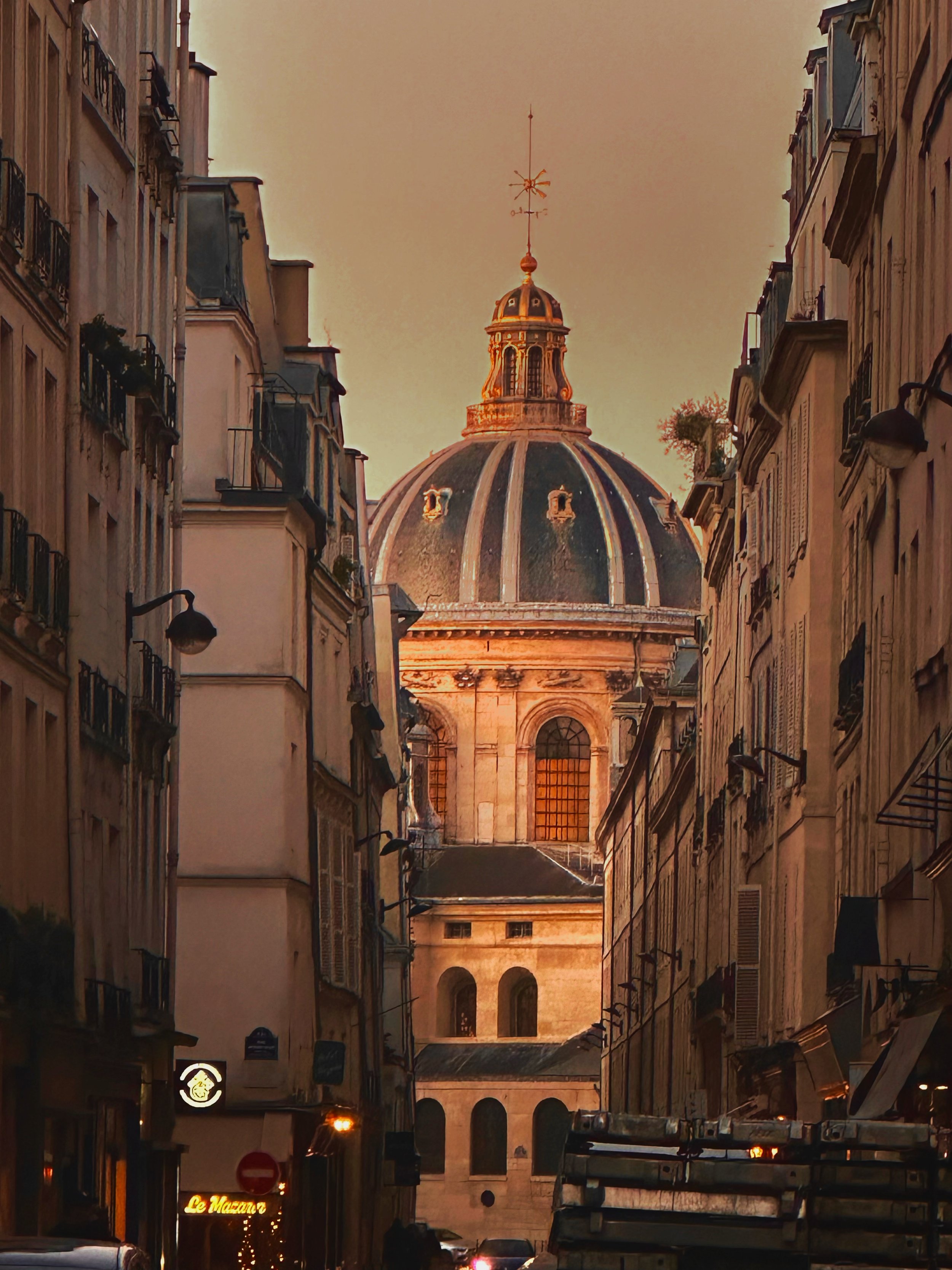 City street view at sunset with tall buildings on both sides and a large domed church in the background. The street has street lamps and a sign that reads 'Le Mazarin'.