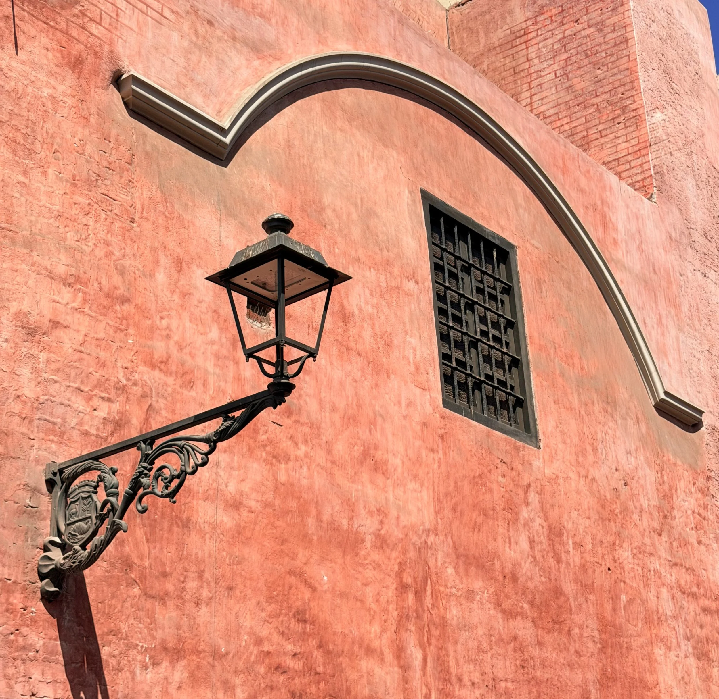 Wrought-iron street lamp against a curved terracotta wall with a small barred window