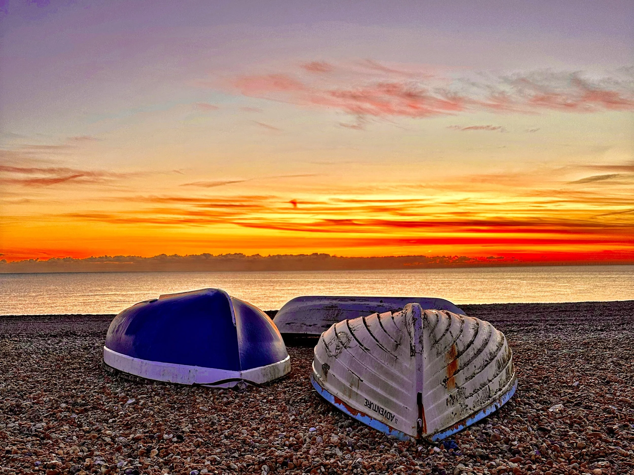 Boats upside down on a pebbled beach during sunrise or sunset with a colorful sky and calm water in the background.