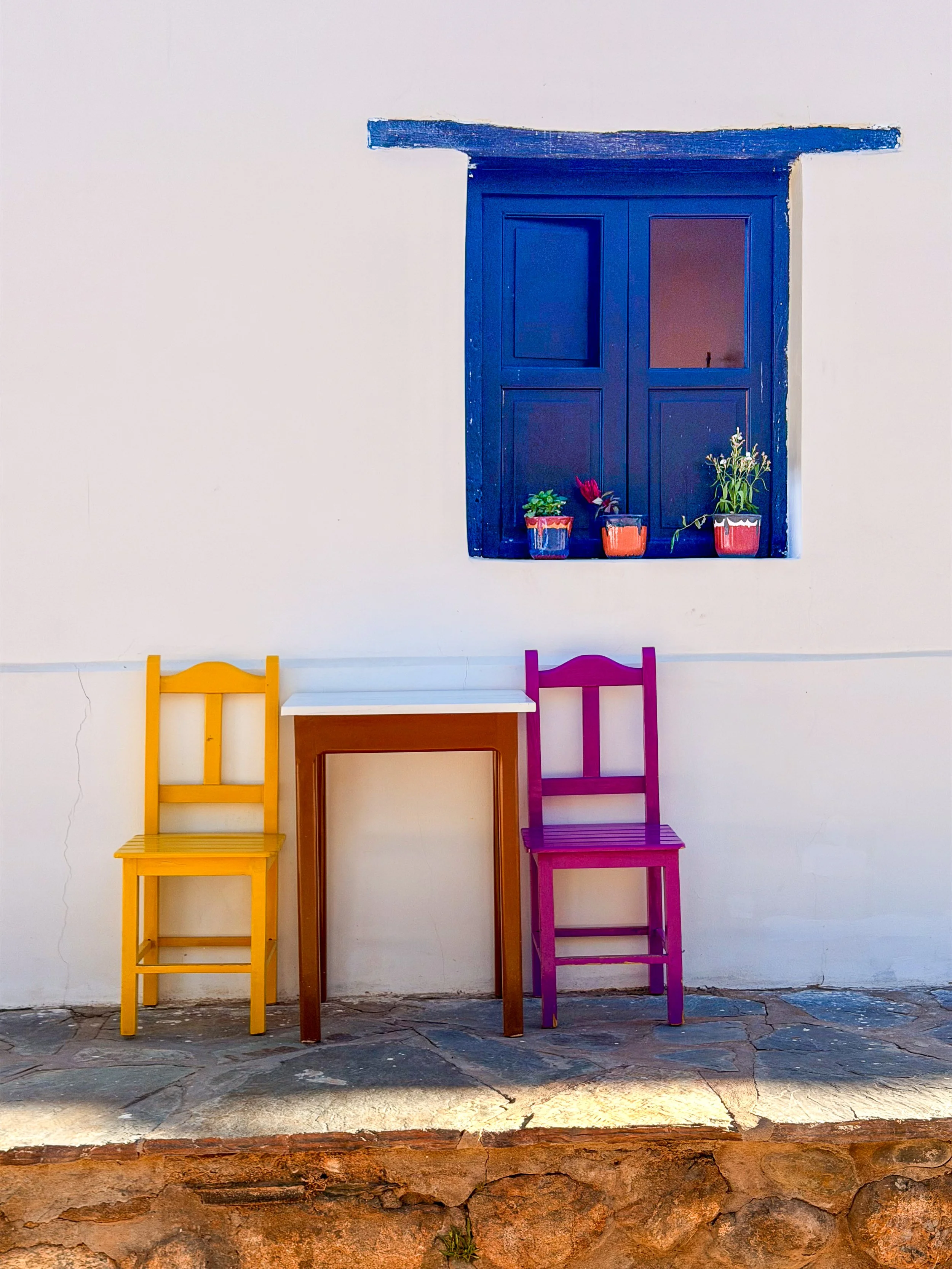 Yellow and magenta chairs flank a table beneath a blue-shuttered window with potted plants