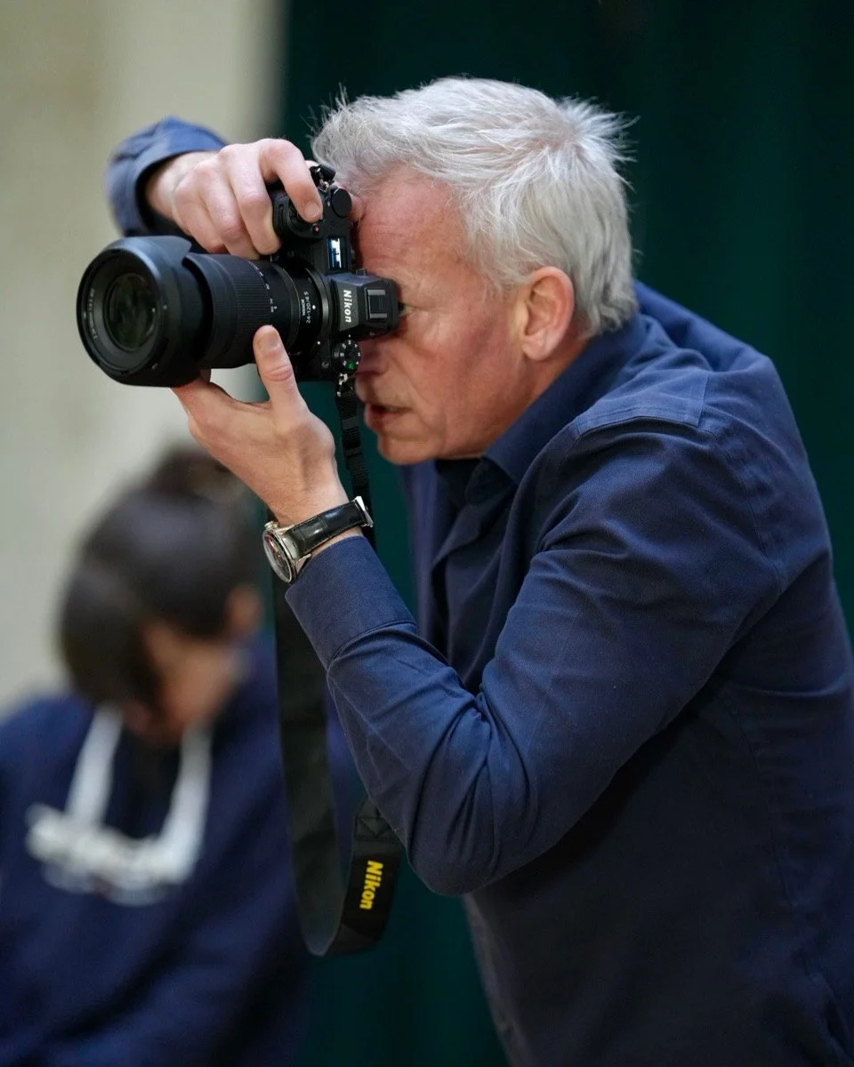 man with gray hair taking a photograph with a Nikon DSLR camera. He is wearing a dark blue shirt and a watch. In the background, a person with dark hair is slightly blurred.