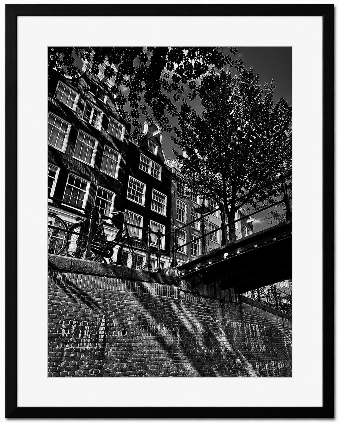 Black and white photo of bicycles parked along a railing along an Amsterdam Canal with tall buildings and trees in the background.