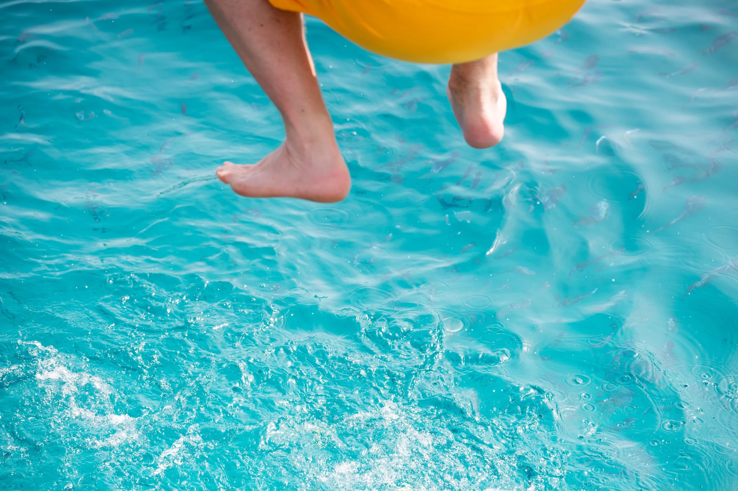 Feet dangling above shimmering turquoise pool water beside a yellow inflatable float