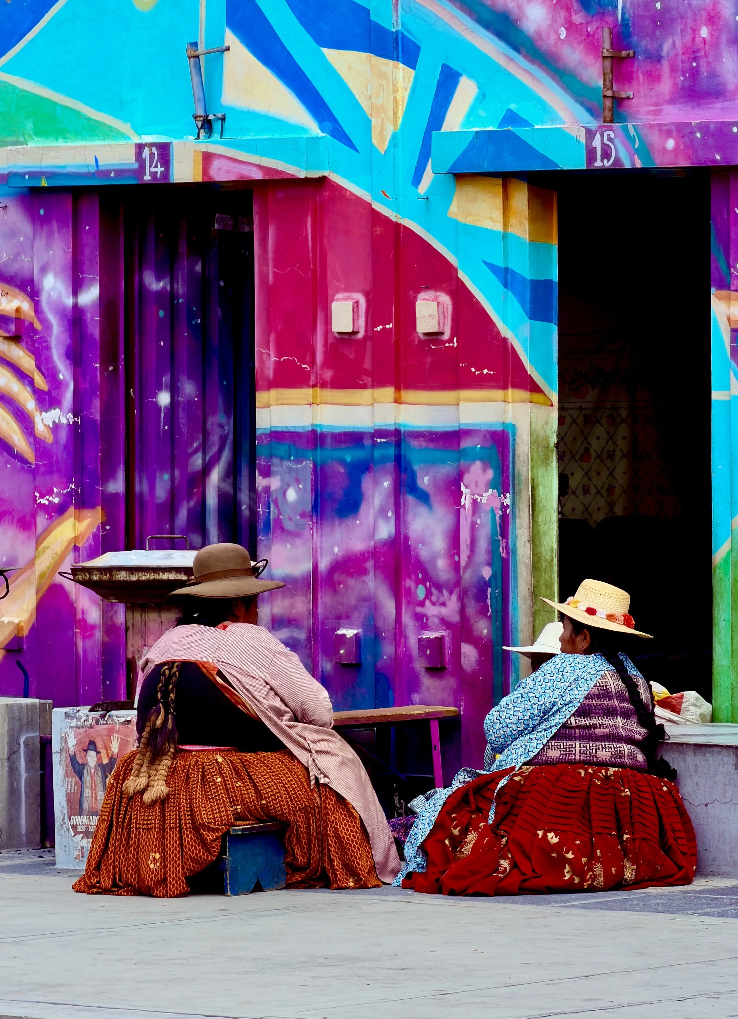 Two women in traditional Bolivian dress and bowler hats sitting before a vivid mural wall