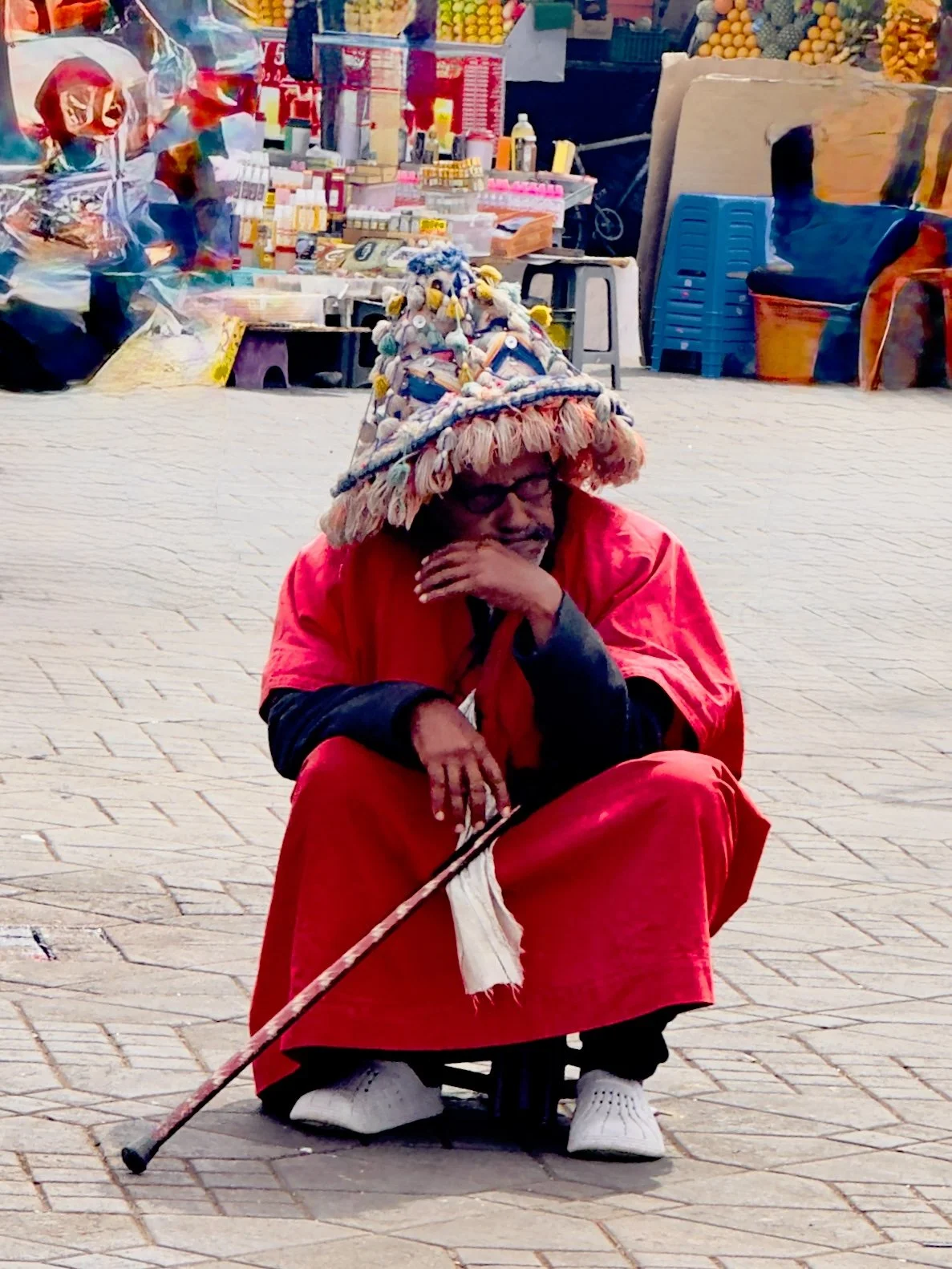 Elderly person in a bright red robe and fringed hat resting on a cane near a market stall