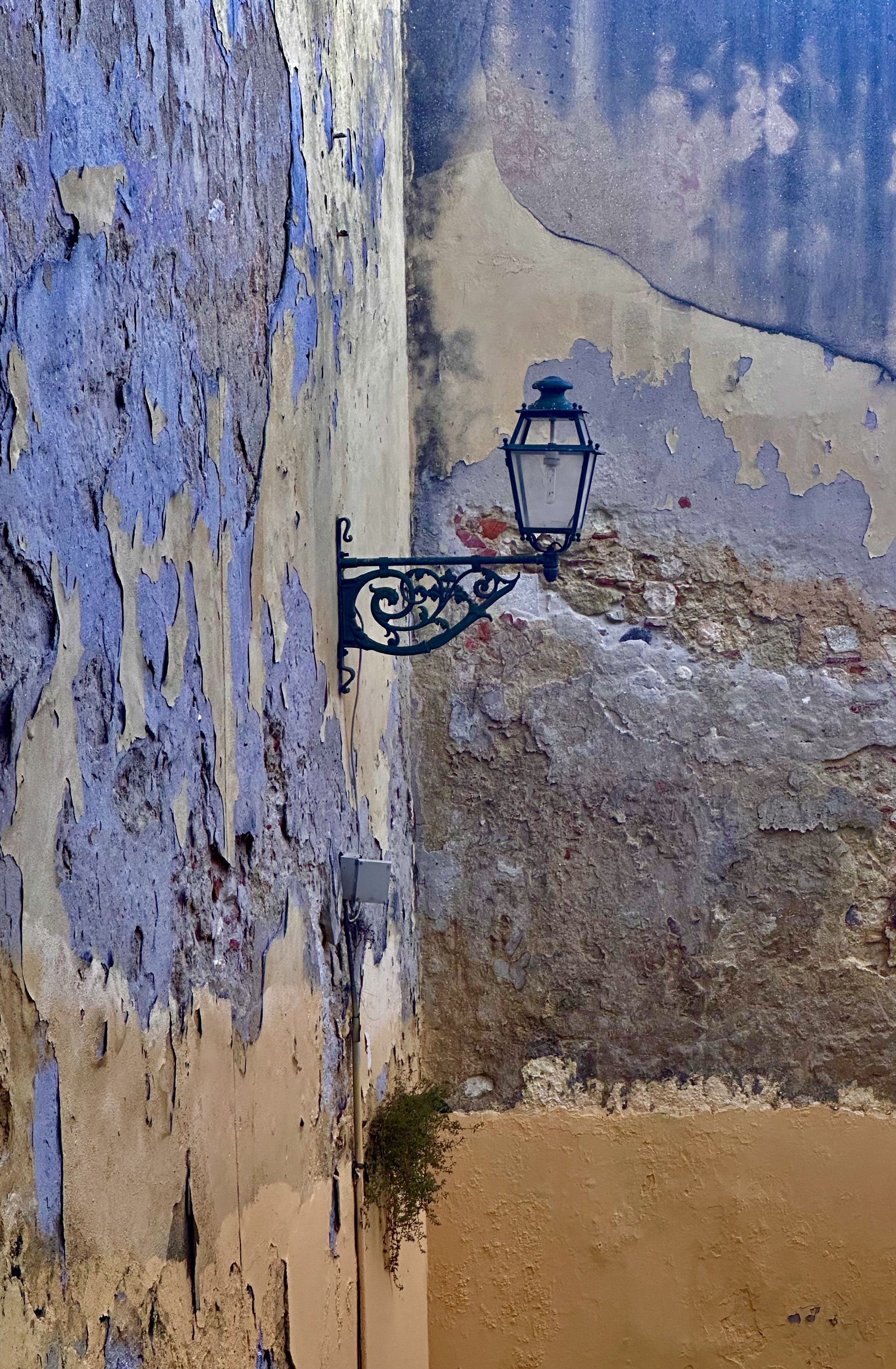 An old, weathered outdoor wall with peeling paint and cracks, featuring a black wrought iron lantern attached to the wall.