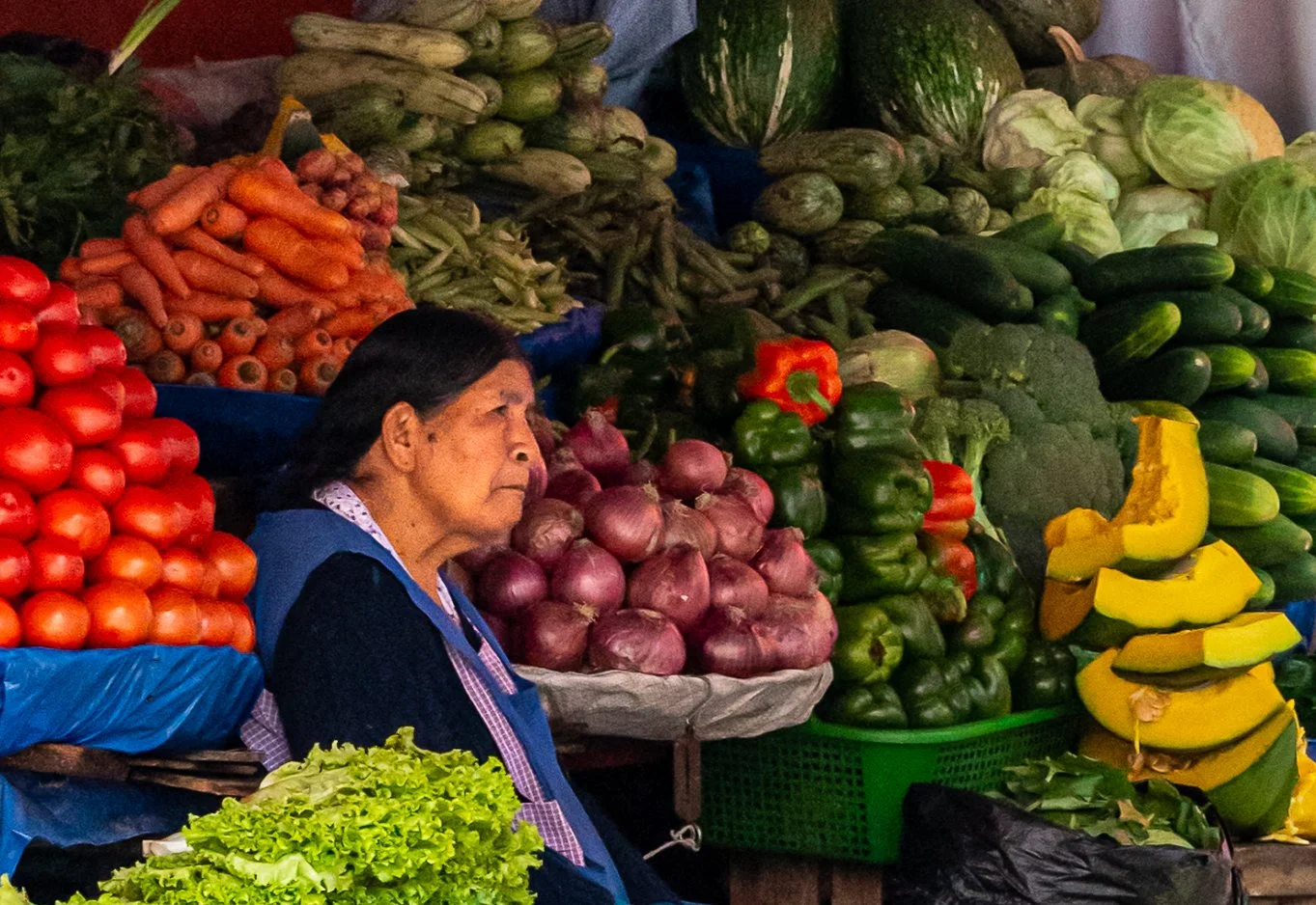 Vendor surrounded by stacks of fresh produce at a colorful vegetable market