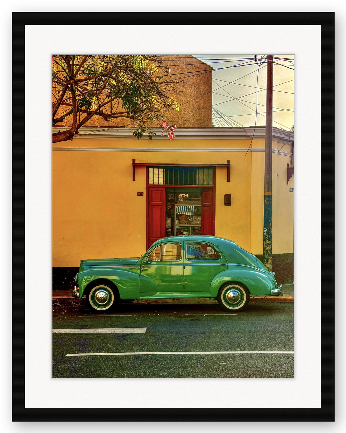 A vintage green car parked in front of a yellow building with a red door, a tree with pink flowers, and utility poles against a partly cloudy sky.