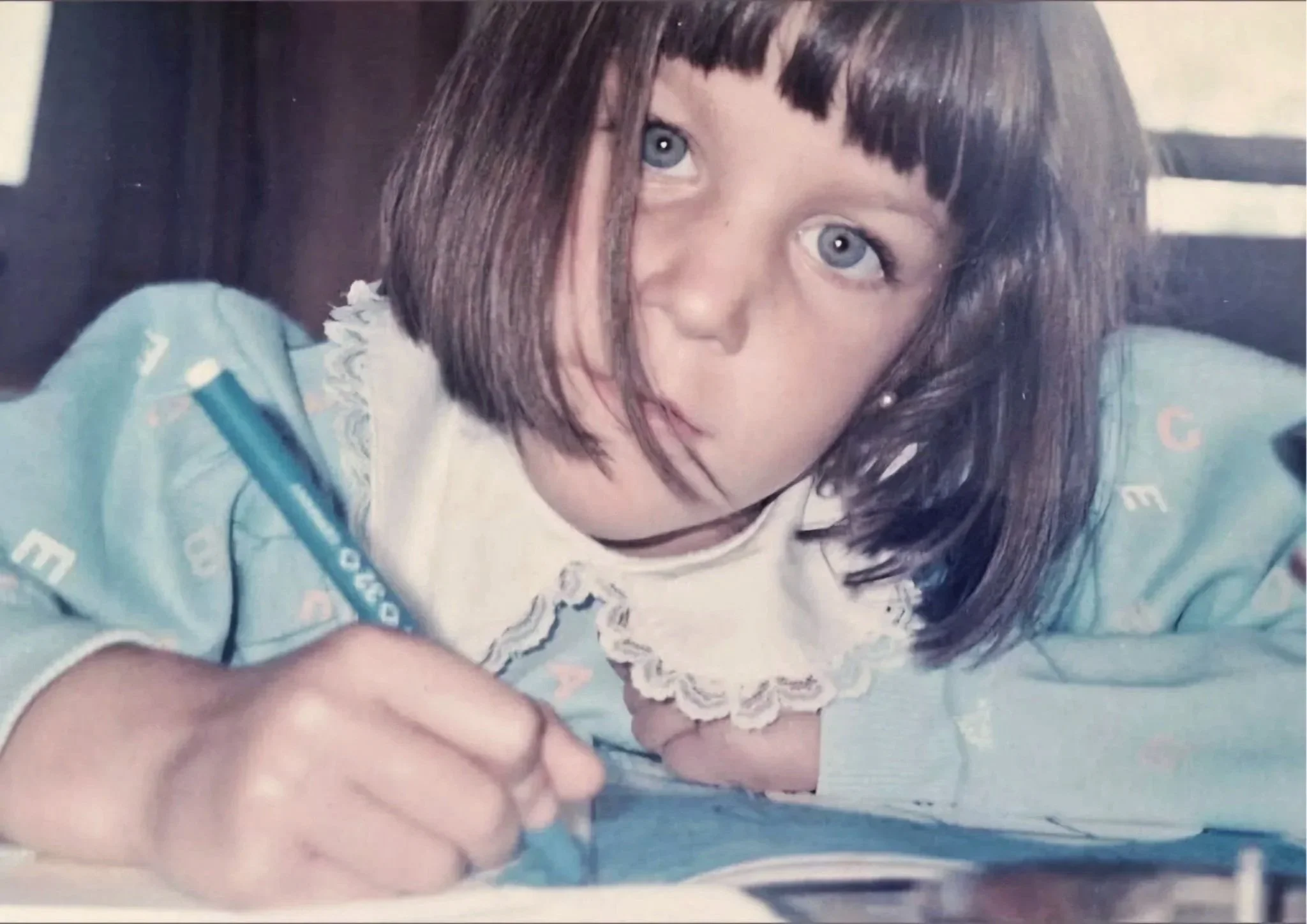 A photograph of Leticia Scottini, the Artist, when she was a young girl with short brown hair and blue eyes, drawing with a pen and looking upward.