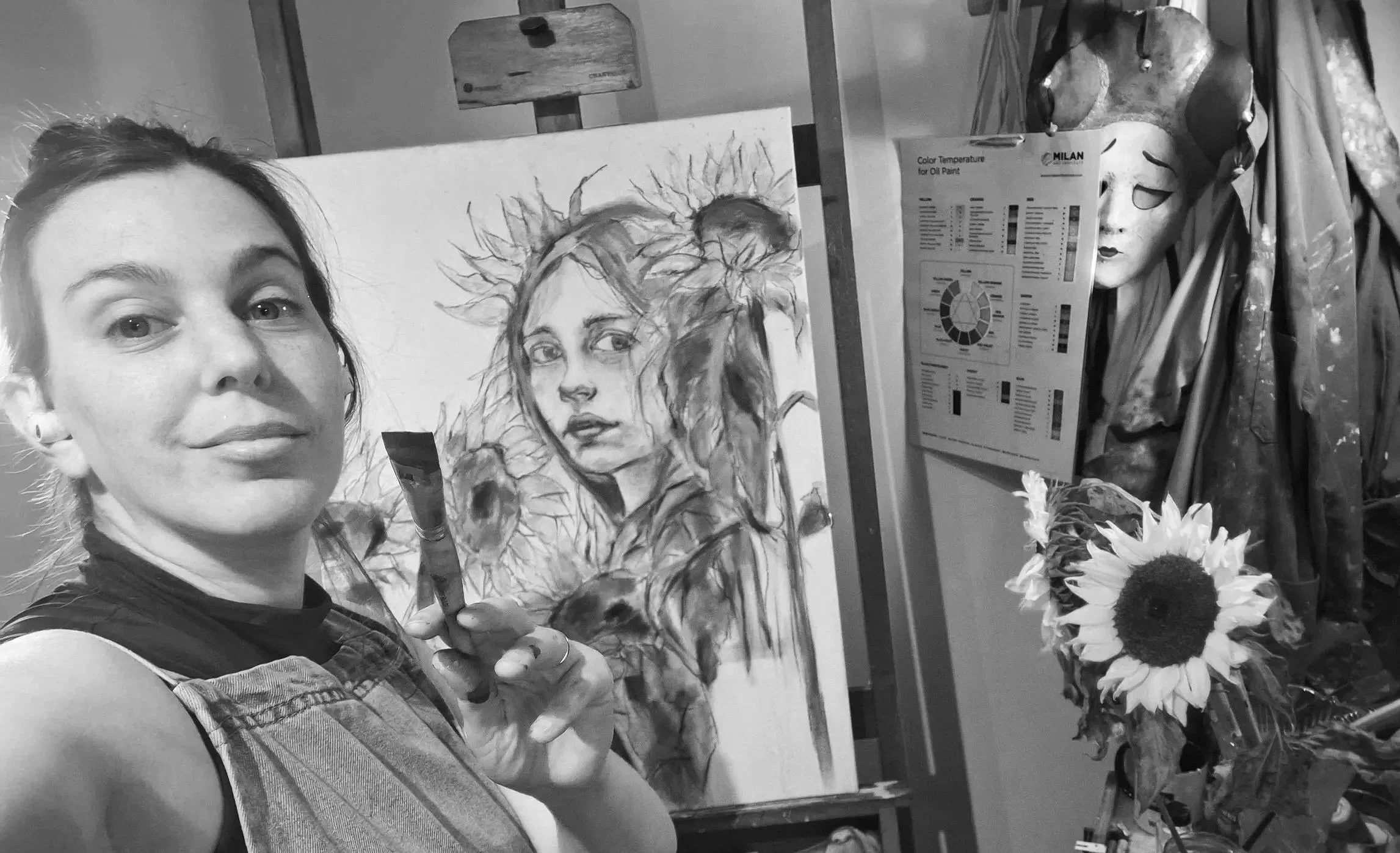 The Artist, Leticia Scottini, holding a paintbrush in front of her unfinished painting  "Sunflower Soul", Next to the painting there is a vase of real sunflowers.