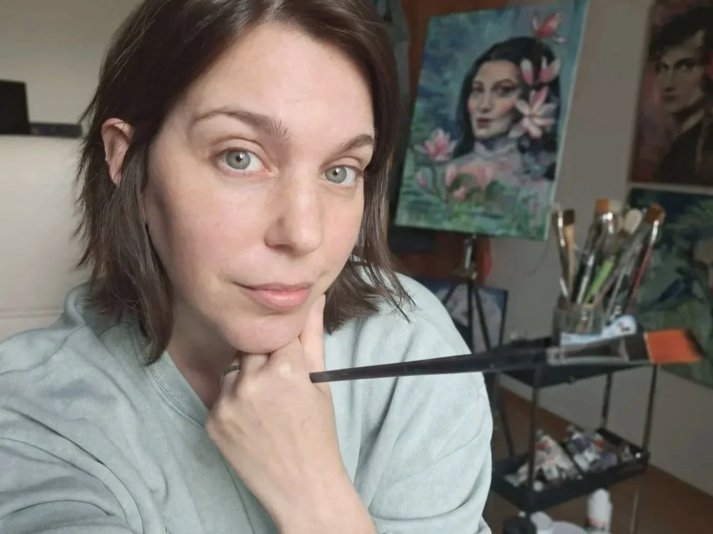 A  photograph of Leticia Scottini, the Artist, short brown hair and light eyes looks at the camera, resting her chin on her hand with a paintbrush, in an art studio with canvases and paint supplies in the background.