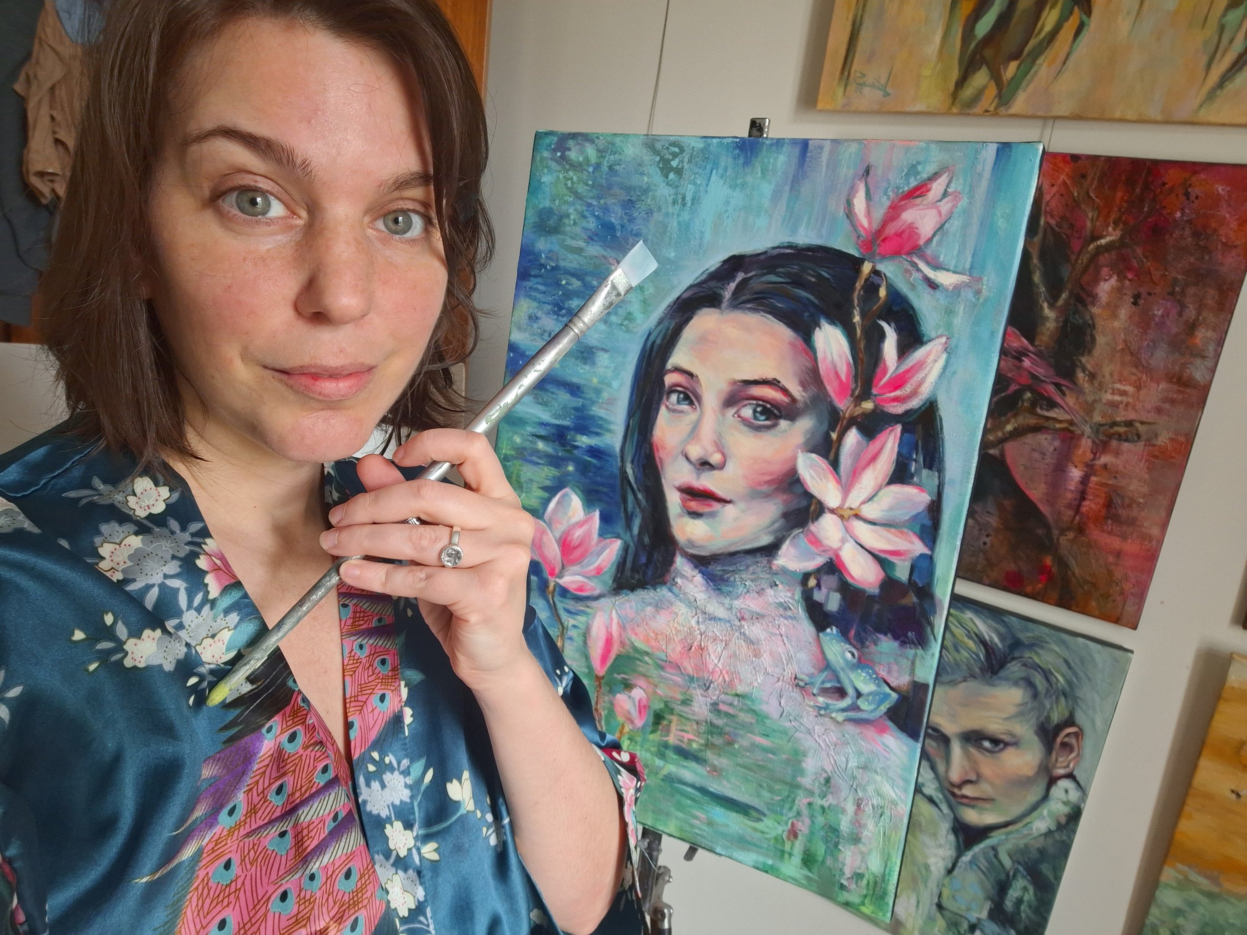 Leticia Scottini, the Artist, holding a paintbrush in front of a colorful portrait painting of a woman with dark hair and pink flowers, emerging from an abstract background that looks like water, this is her painting called "Silently emerging"