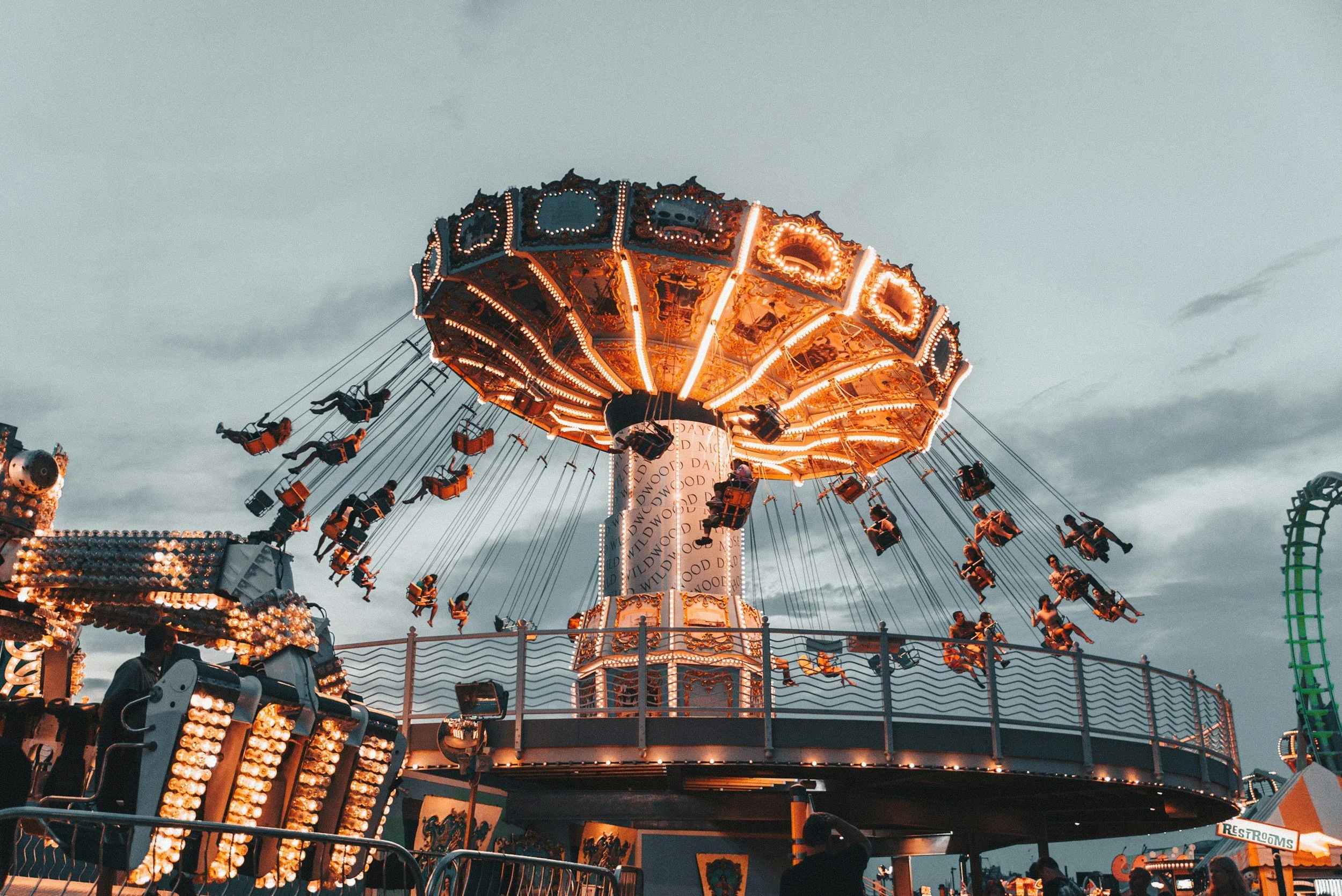 A carousel ride at a fair with swing seats filled with riders, illuminated with bright orange lights, against a dusk sky.