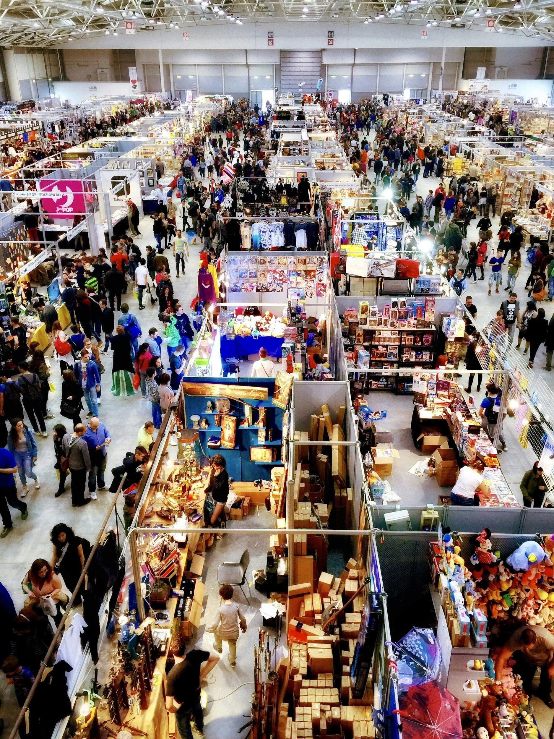 Wide view of a crowded indoor market or convention with numerous vendor booths and visitors browsing, some colorful merchandise on display.
