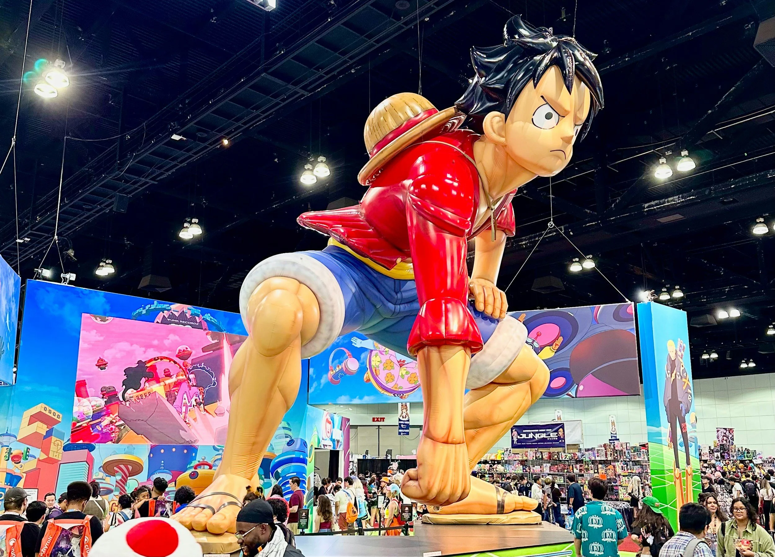 Large statue of Monkey D. Luffy from the anime One Piece, kneeling with a serious expression, in a crowded indoor event with colorful banners and fans.