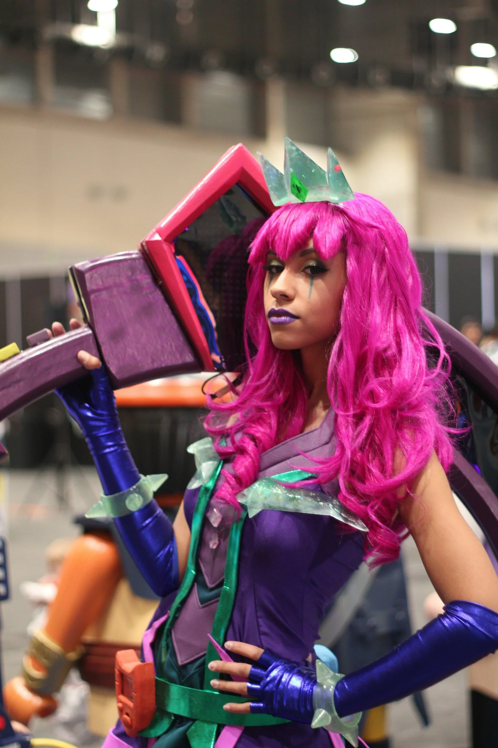 A woman dressed in a colorful, sci-fi inspired costume with pink hair, purple lipstick, and teal makeup with a tear streak. She wears a crown made of translucent, green shards. The costume features purple and green armor-like pieces with shiny blue gloves, and she poses with one hand on her hip.