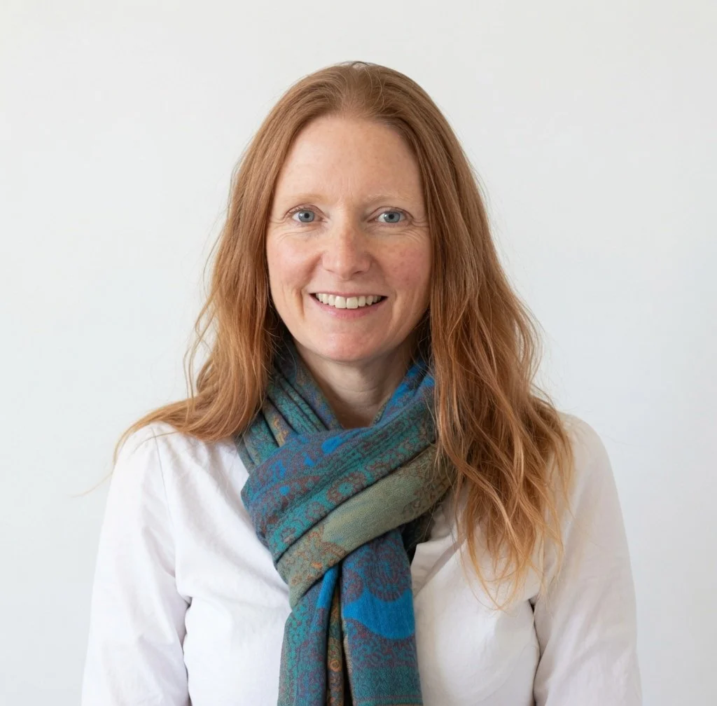 Gillian Mickelson pictured as a smiling woman with long red hair wearing a white shirt and a colorful scarf, standing against a plain white background.