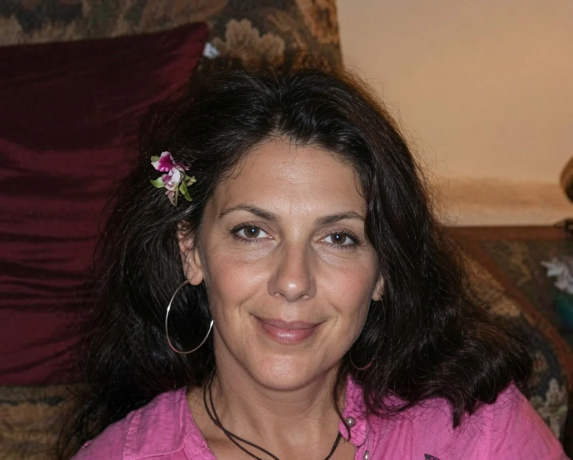 Joyce Roetter pictured with dark, curly hair wearing a pink shirt and hoop earrings, smiling, with a flower tucked behind her ear, sitting on a couch with a decorative pillow.