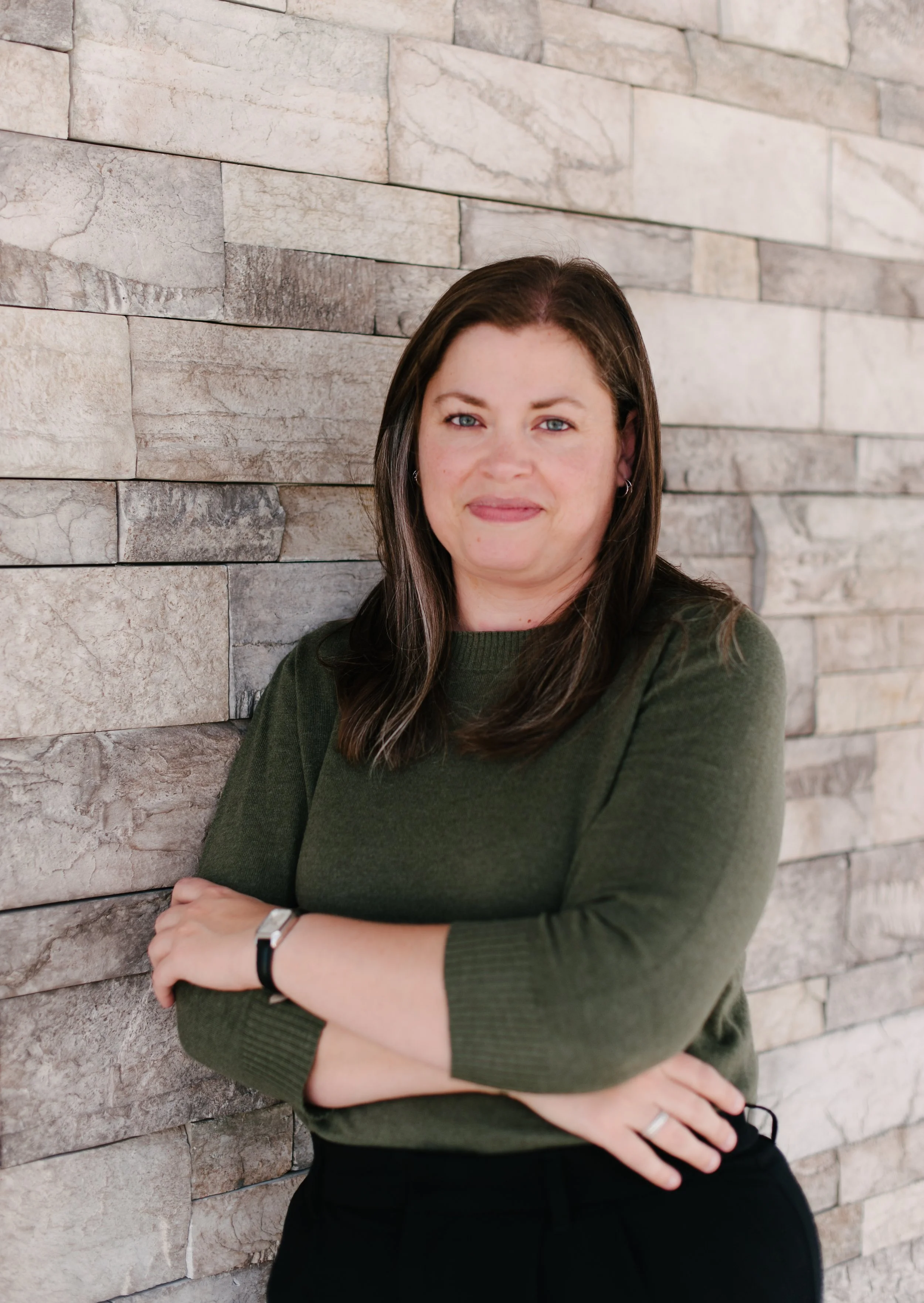 A woman with brown hair and blue eyes standing against a beige stone wall, wearing a green long-sleeve shirt and black pants, with arms crossed and a slight smile.