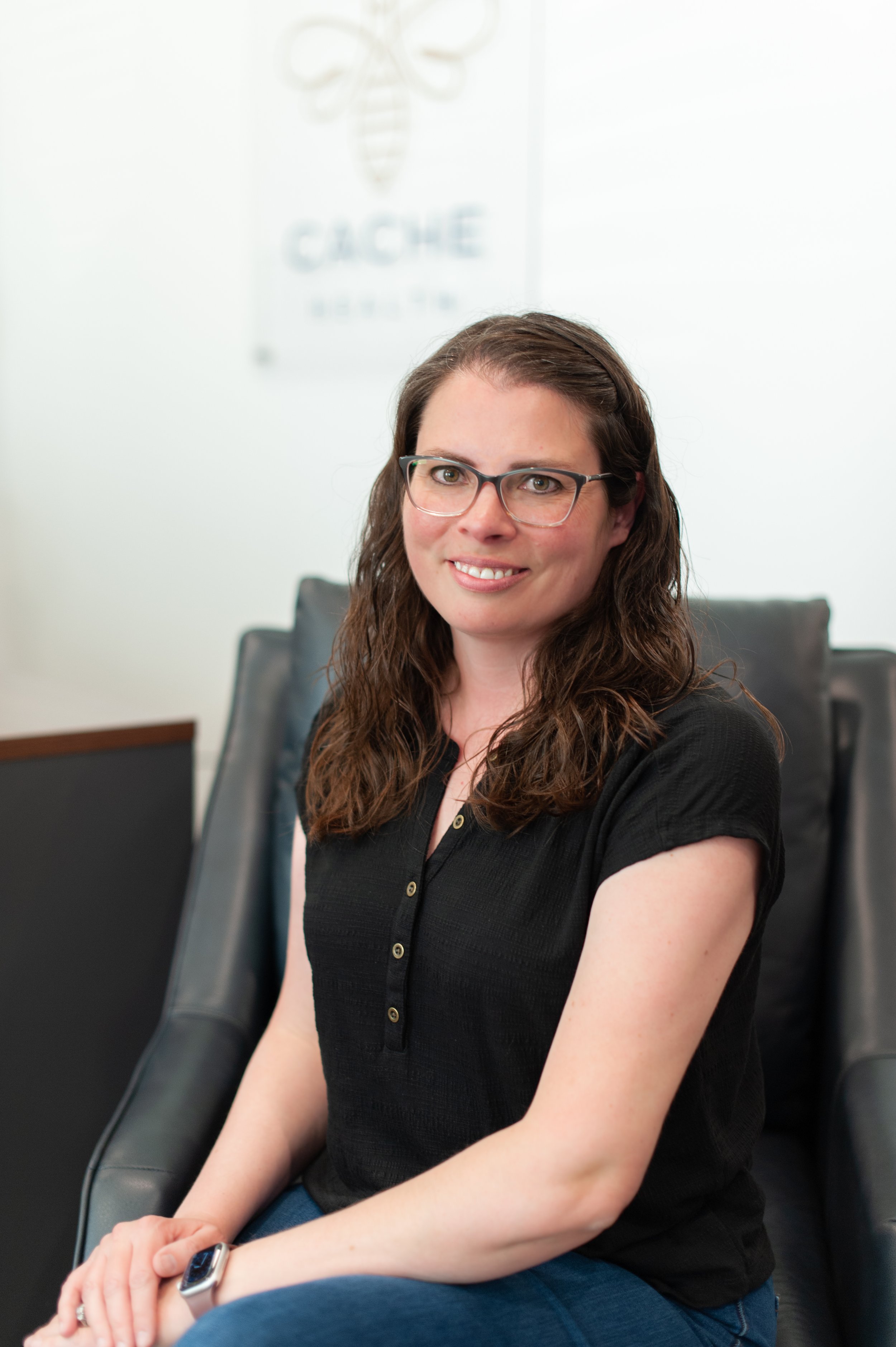 A woman with long, curly brown hair and glasses sitting in a black office chair, smiling at the camera in an office setting. She is wearing a black button-up shirt and a smartwatch on her left wrist.