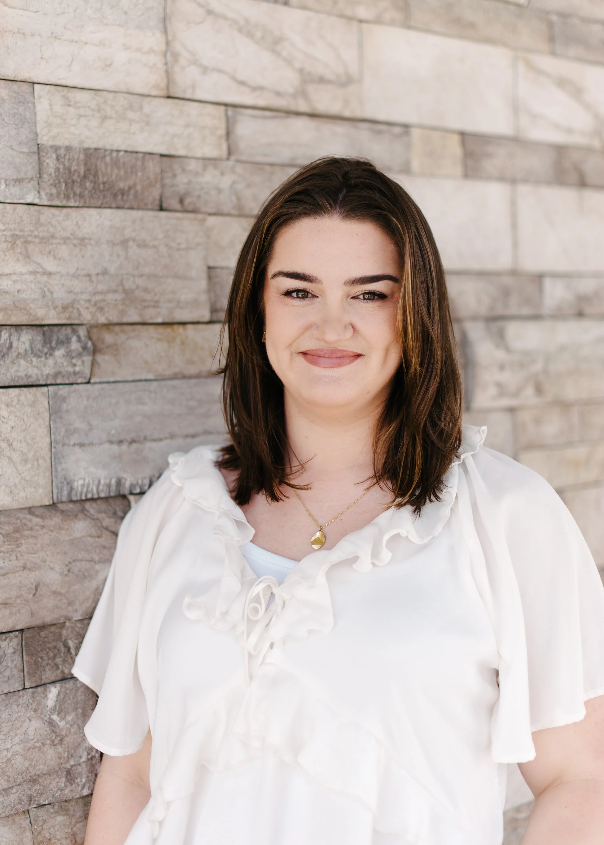 A woman with shoulder-length brown hair, wearing a white blouse with ruffled sleeves and a gold necklace, smiling while standing against a beige stone brick wall.