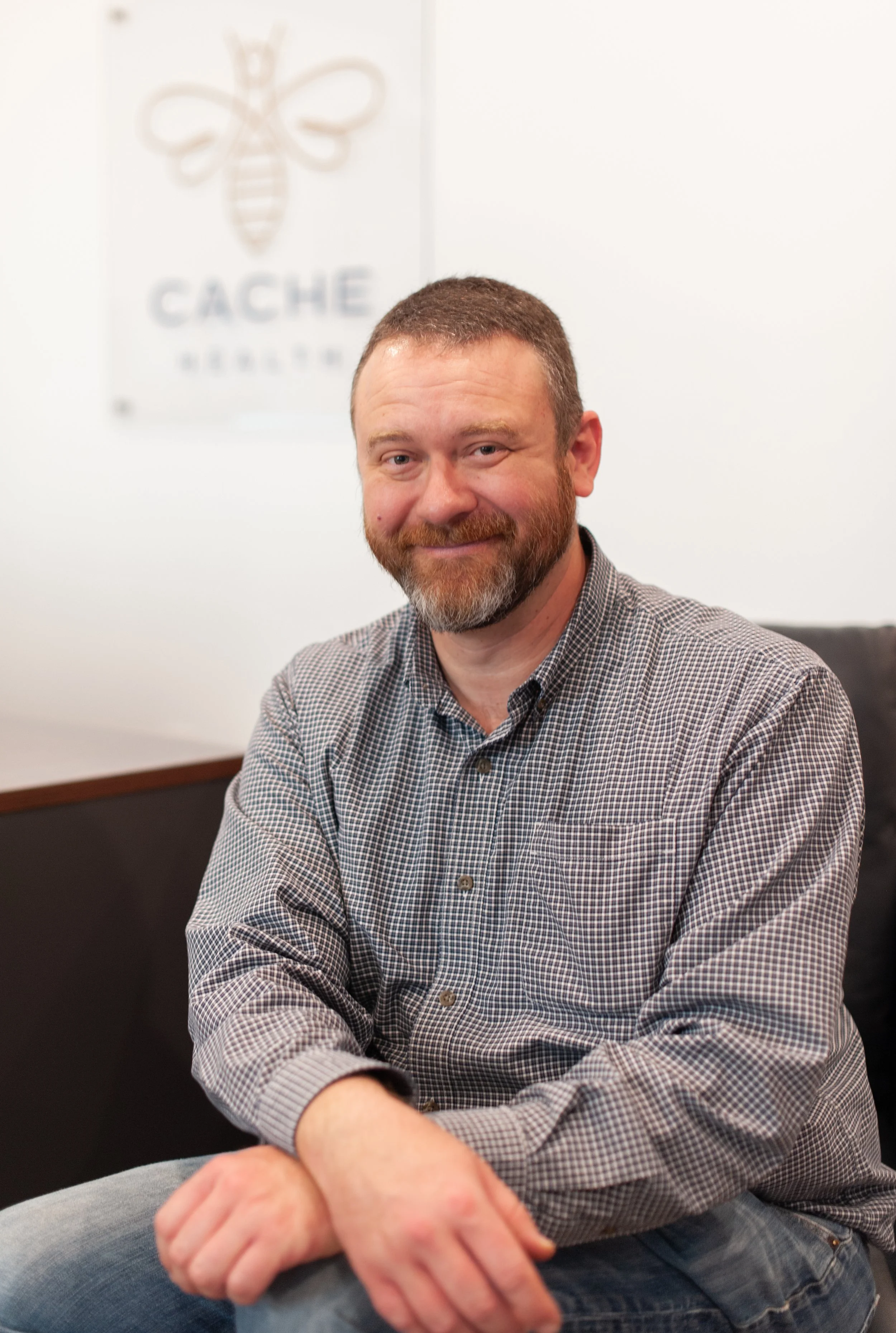 A man with a beard and short hair smiling, sitting in front of a wall with a blurred logo that includes a bee and the words "Cache Honey".
