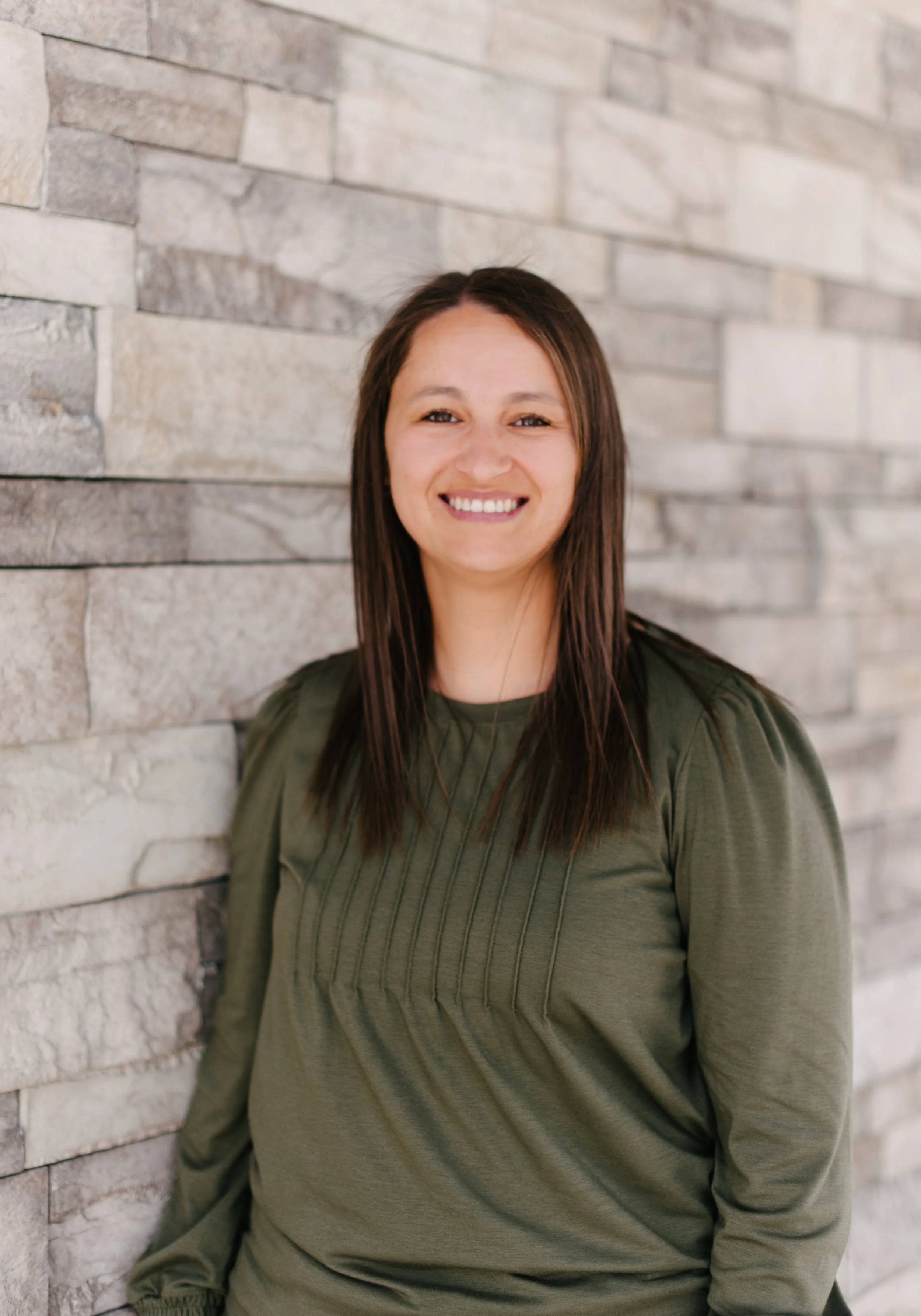 A smiling woman with long brown hair, wearing a dark green ribbed sweater, standing against a textured stone wall.