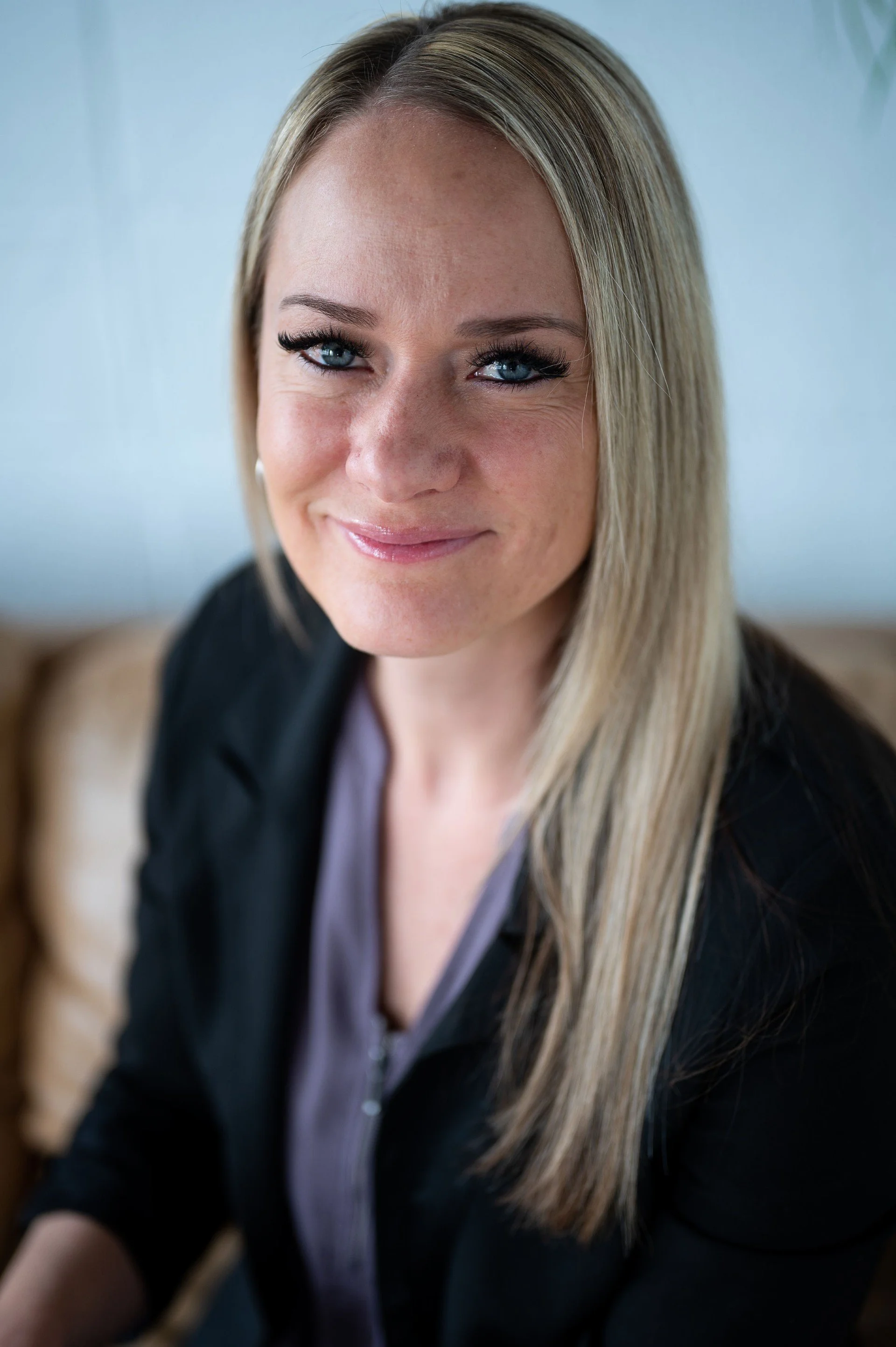 Close-up of a smiling woman with blonde hair and blue eyes, wearing a black jacket and a light gray top.