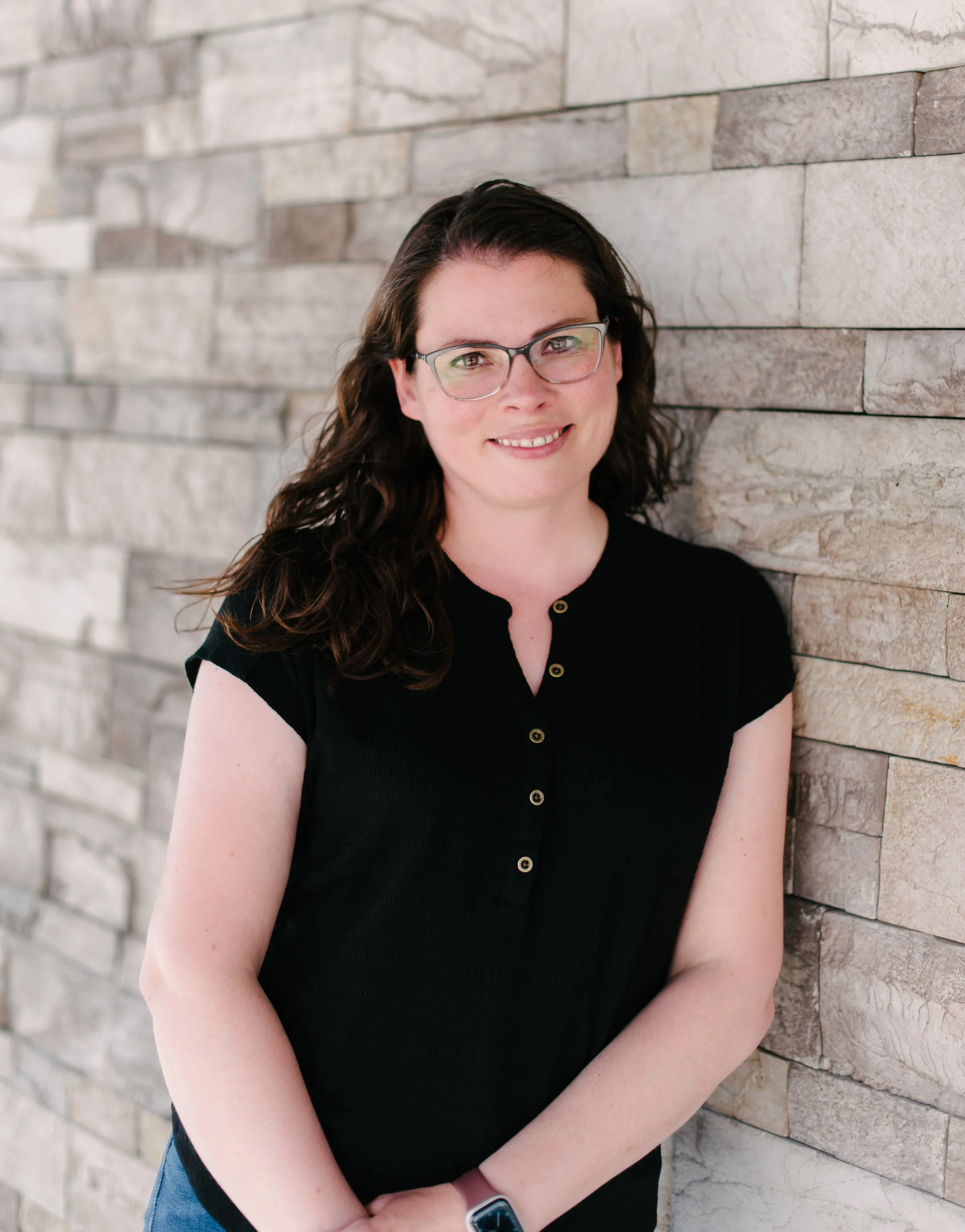 A woman with long dark curly hair and glasses, smiling, wearing a black shirt and standing against a stone wall.