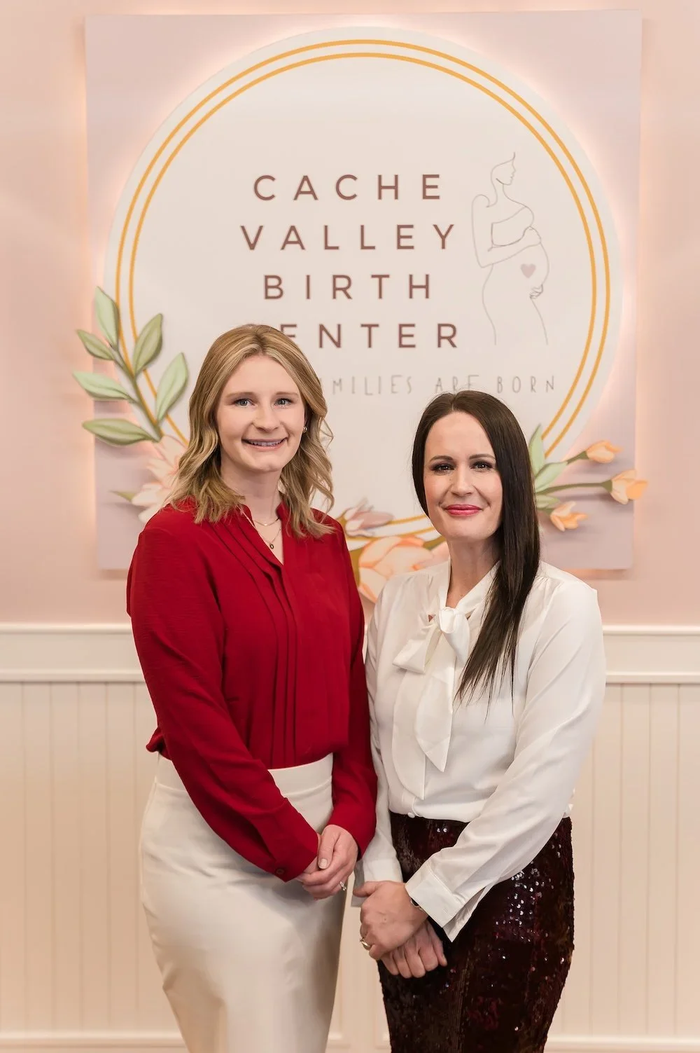 Two women standing in front of a sign that reads "Cache Valley Birth Center, Miles Are Born." One woman has blond hair and is wearing a red blouse and white skirt. The other woman has black hair and is wearing a white blouse with a bow tie and black sequined pants. The background has a pale pink wall with light decor.