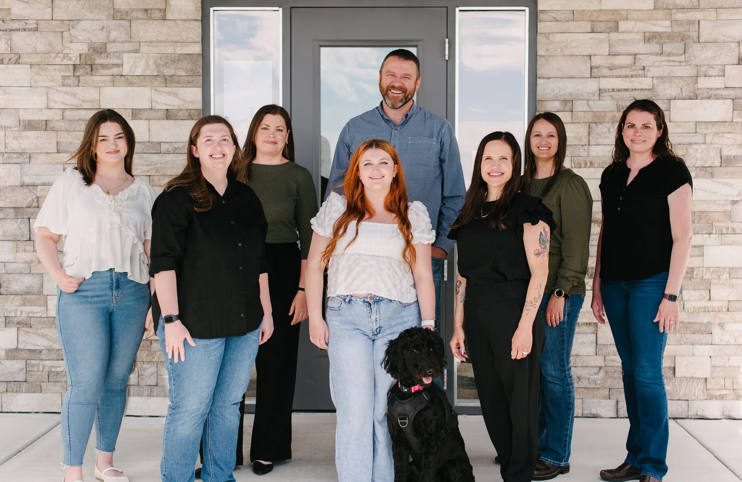 A group of nine people and one dog standing in front of a modern stone wall and glass door, smiling for the camera.