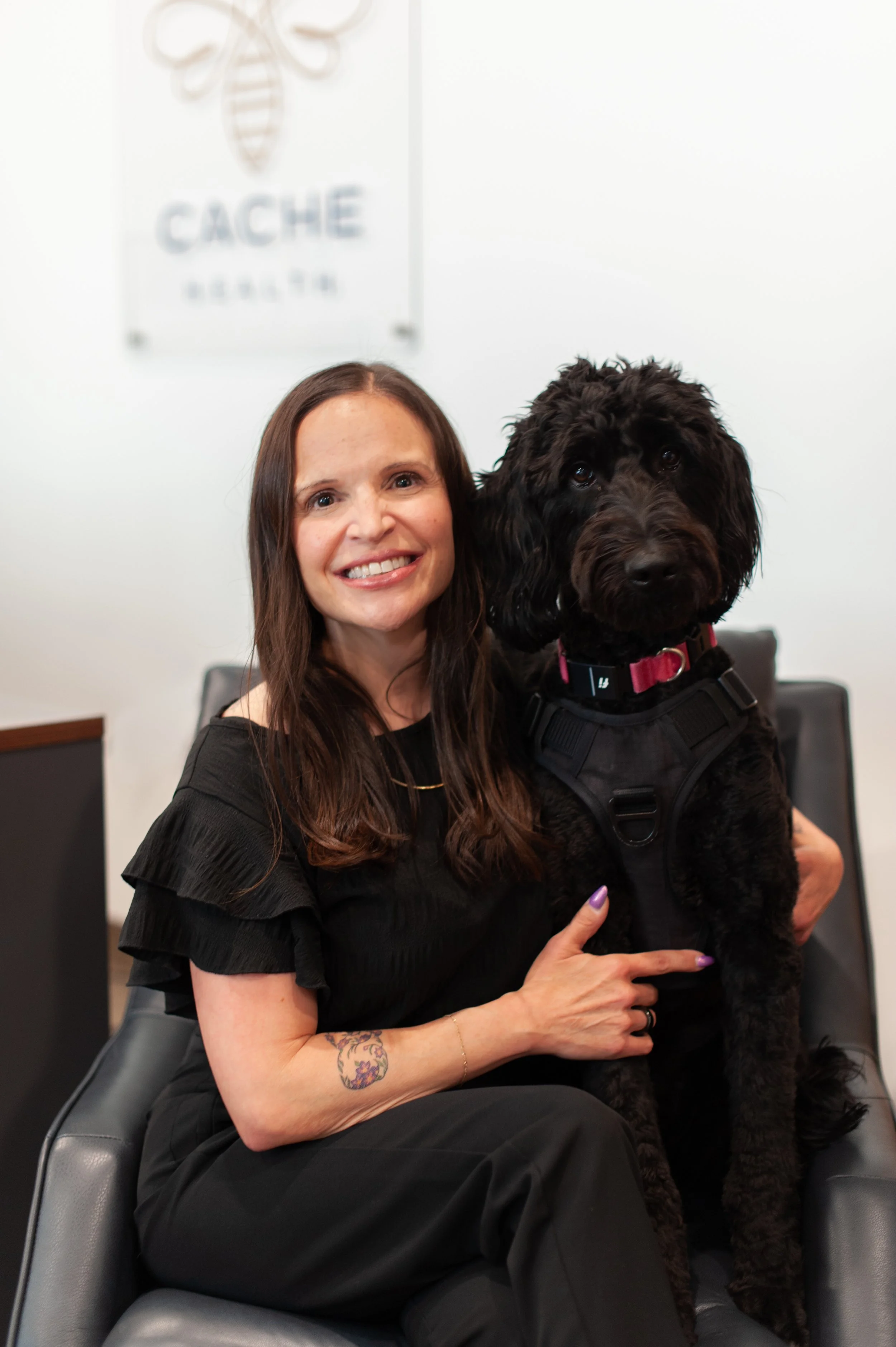 A woman with long dark hair and a tattoo on her left arm, sitting in a gray chair, smiling and holding a large black dog with curly fur and floppy ears, wearing a pink collar and a harness, in an indoor setting with a white wall and a sign that says "CACHÉ" in the background.