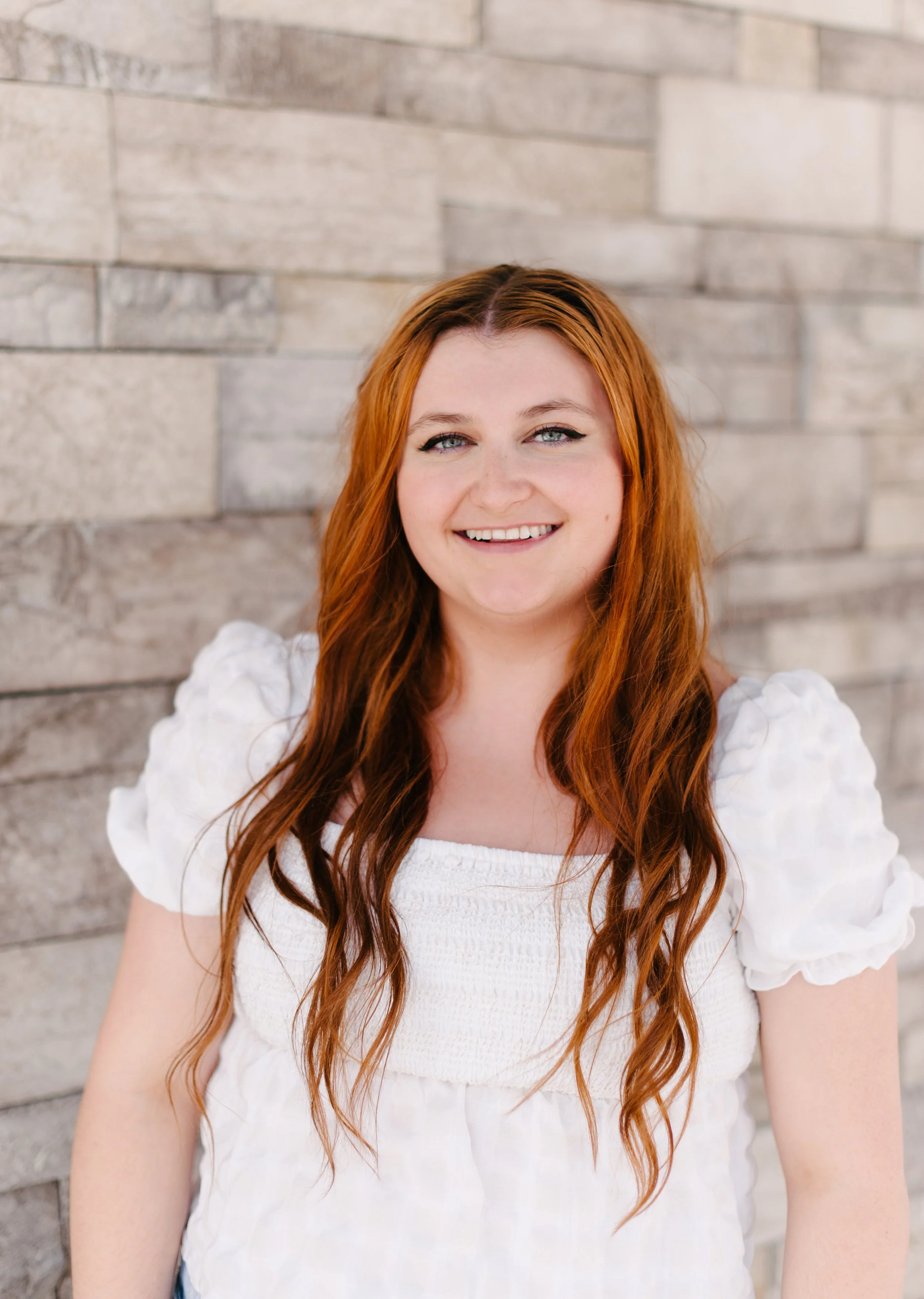 A woman with long red hair and blue eyes smiling at the camera, wearing a white puff-sleeve top, standing against a gray brick wall.