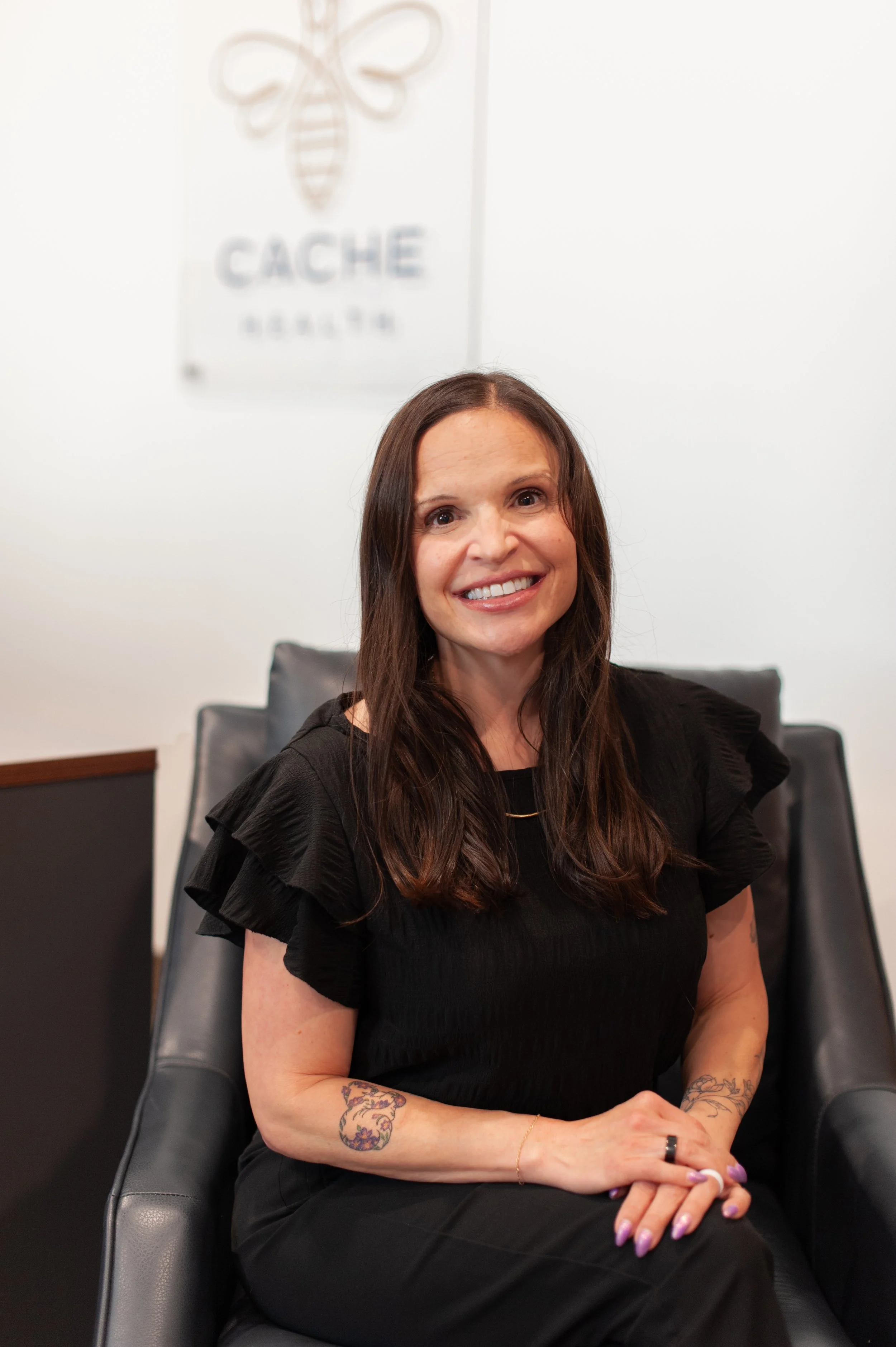 A woman with long brown hair and tattoos on her arms, sitting in a black leather chair, smiling, wearing a black dress with ruffled sleeves and purple nail polish.