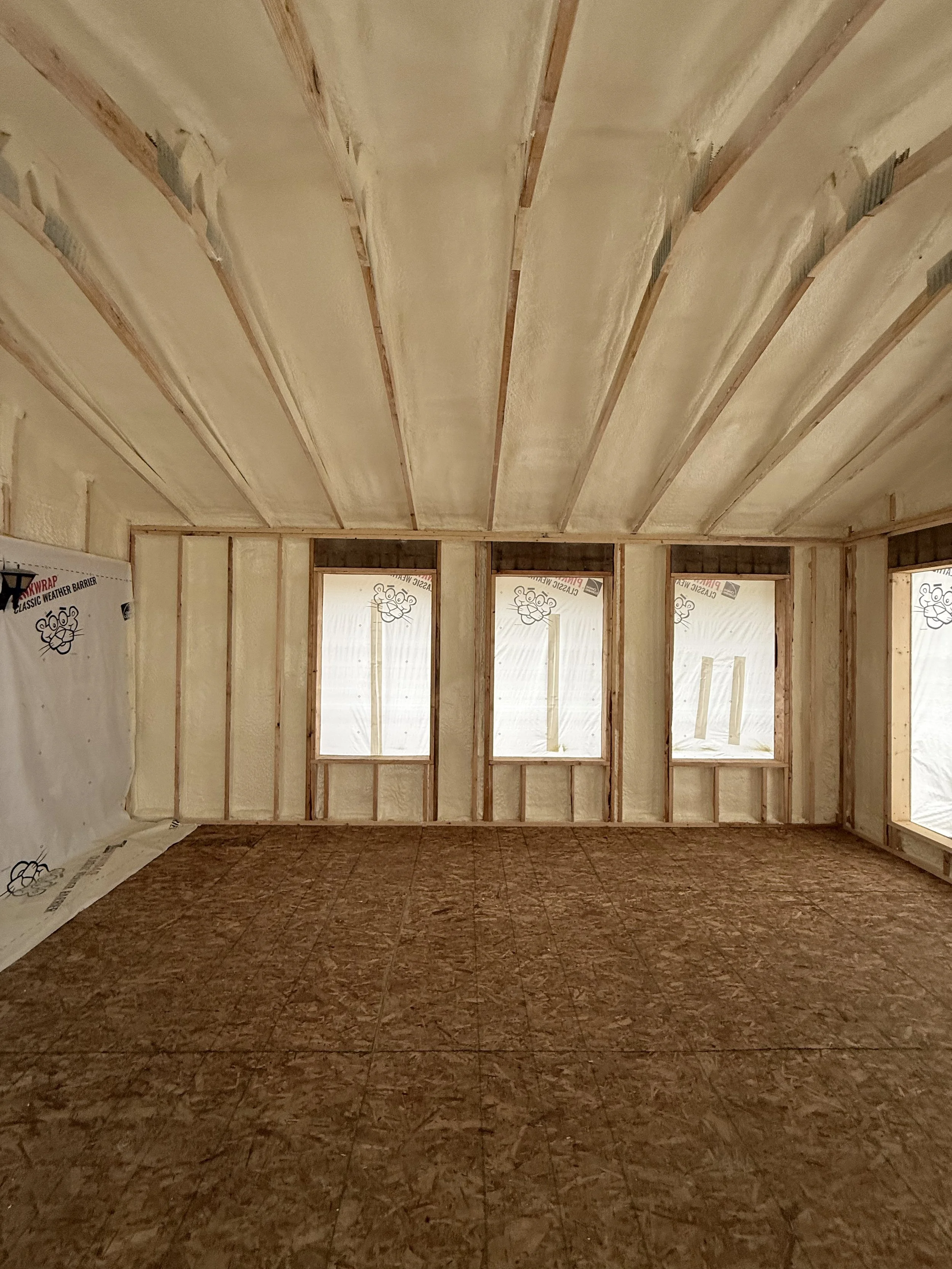 Interior view of a room under construction with exposed wall studs, insulation, and a plywood subfloor.