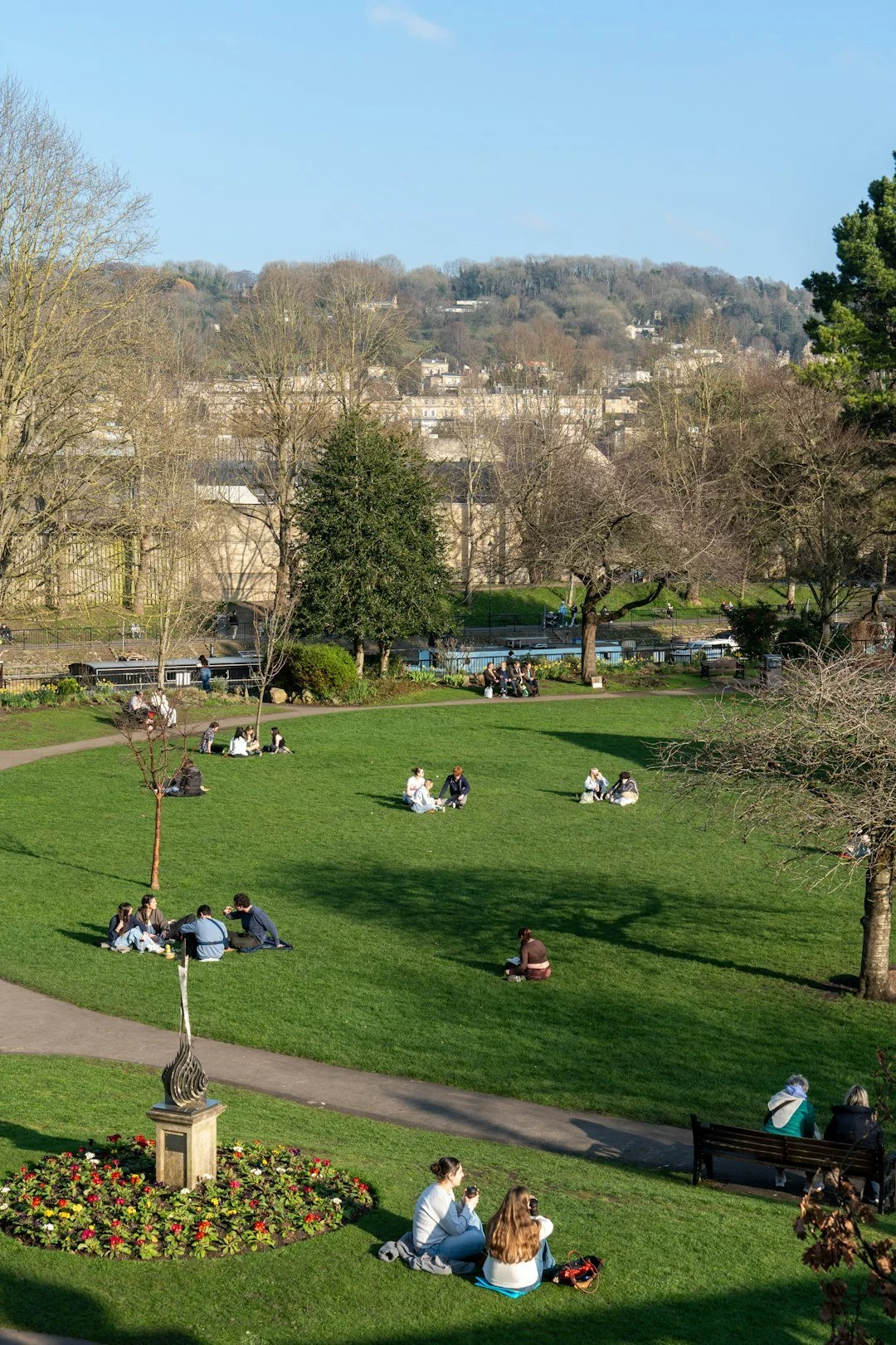 People sitting on the grass in a park on a sunny day, with trees, a flower bed, and a cityscape in the background.