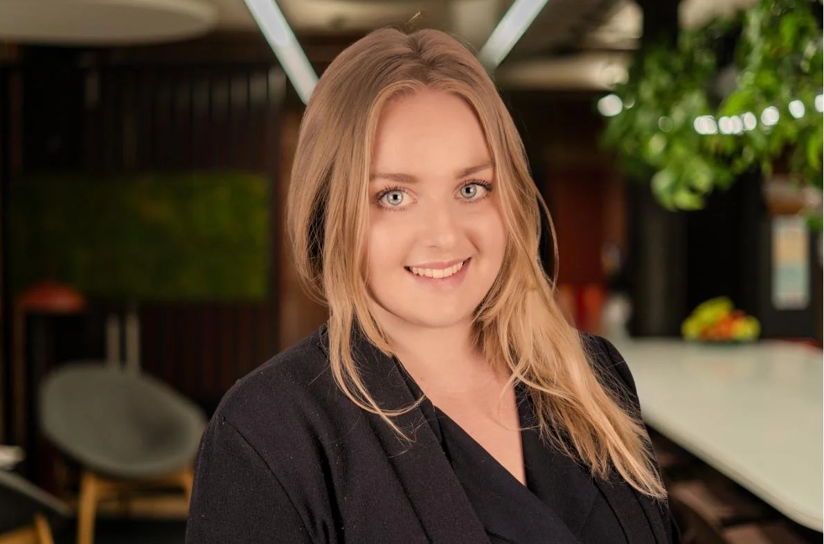 A woman with shoulder-length blonde hair and blue eyes, smiling, in an indoor setting with green plants and modern decor.