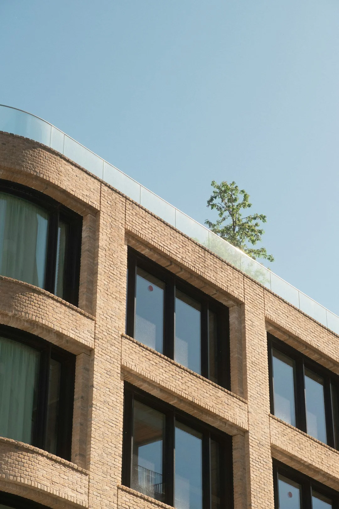 A modern multi-story building with brick exterior, large black-framed windows, and a rooftop with a glass railing and a small tree, under a clear blue sky.