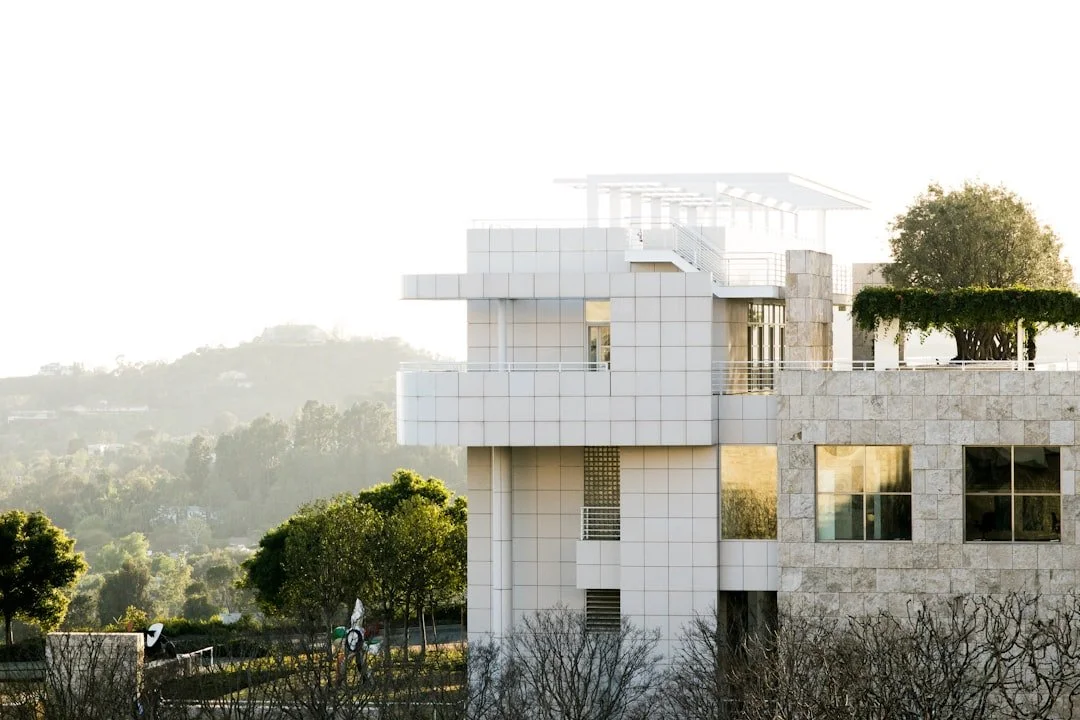 Modern white building with stone accents and fenced terrace, surrounded by trees and hilly landscape