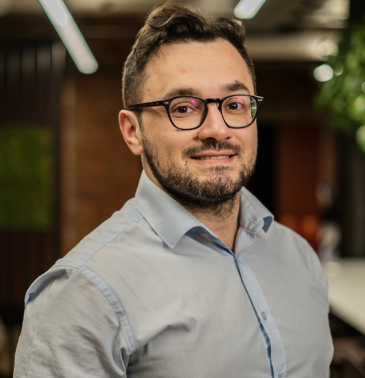 A man with glasses and a beard smiling in an indoor office space.