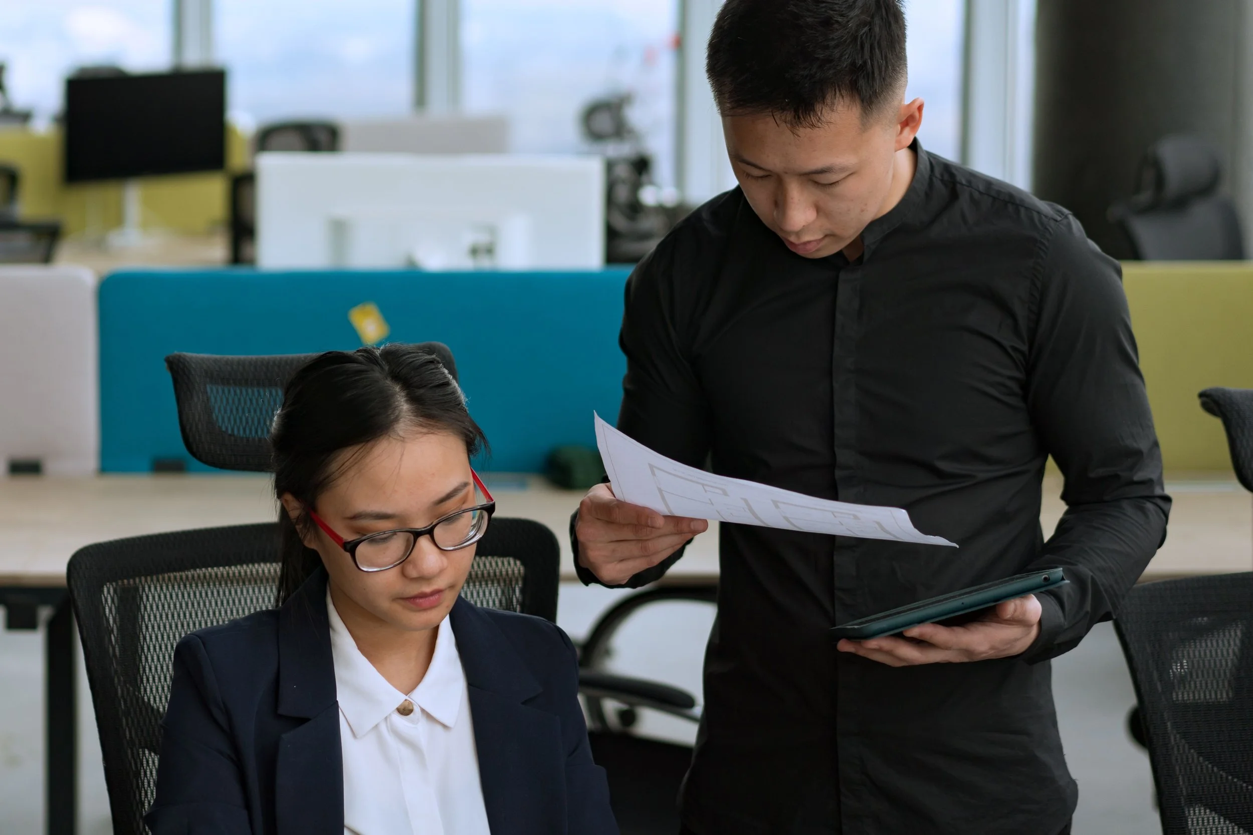 A young man and woman in a modern office, with the man holding a tablet and paper, and the woman sitting at a desk wearing glasses and a business suit.