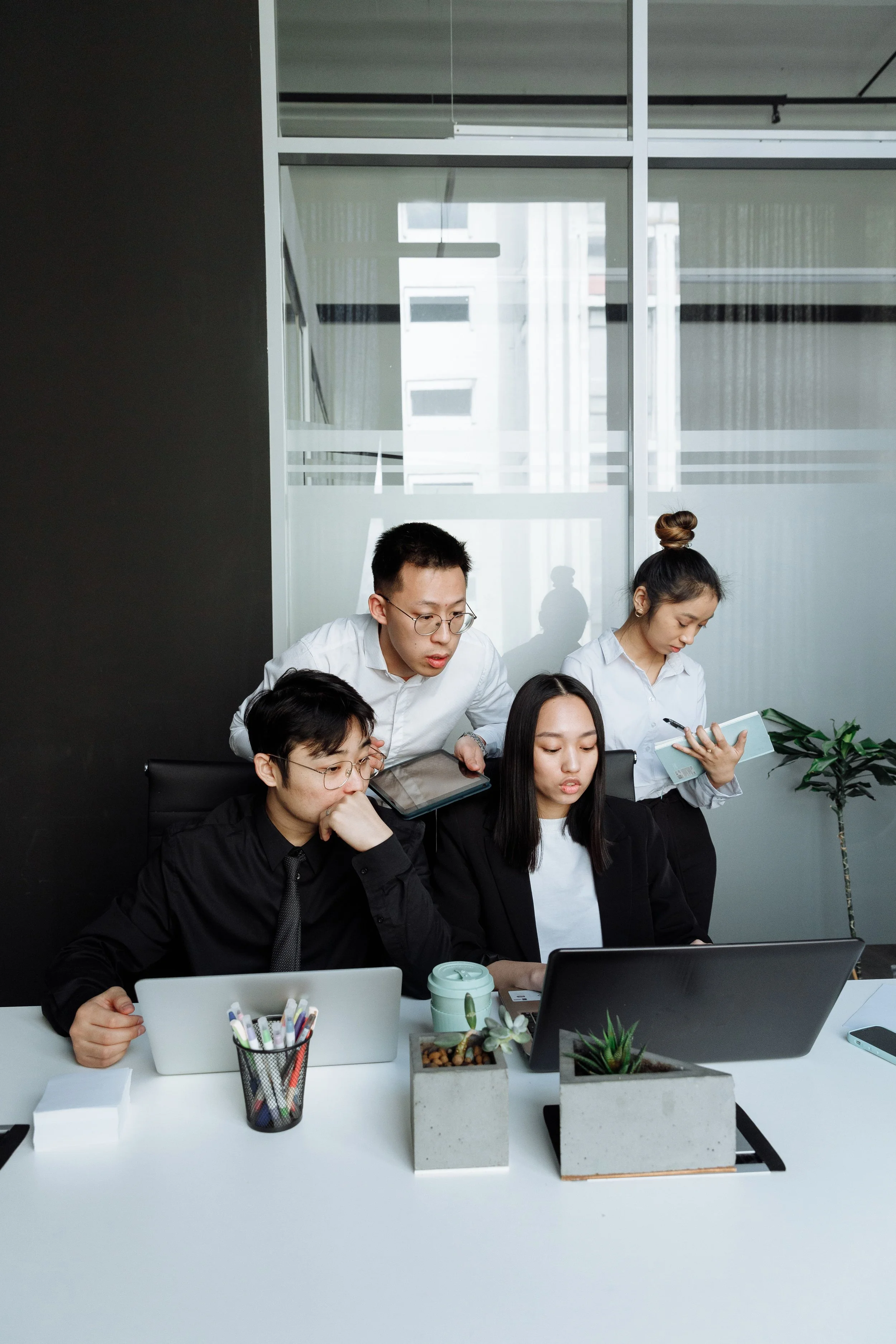 Office meeting with five Asian professionals working at a white desk with laptops, notebooks, plants, and office supplies, in a modern conference room with glass walls and city view.
