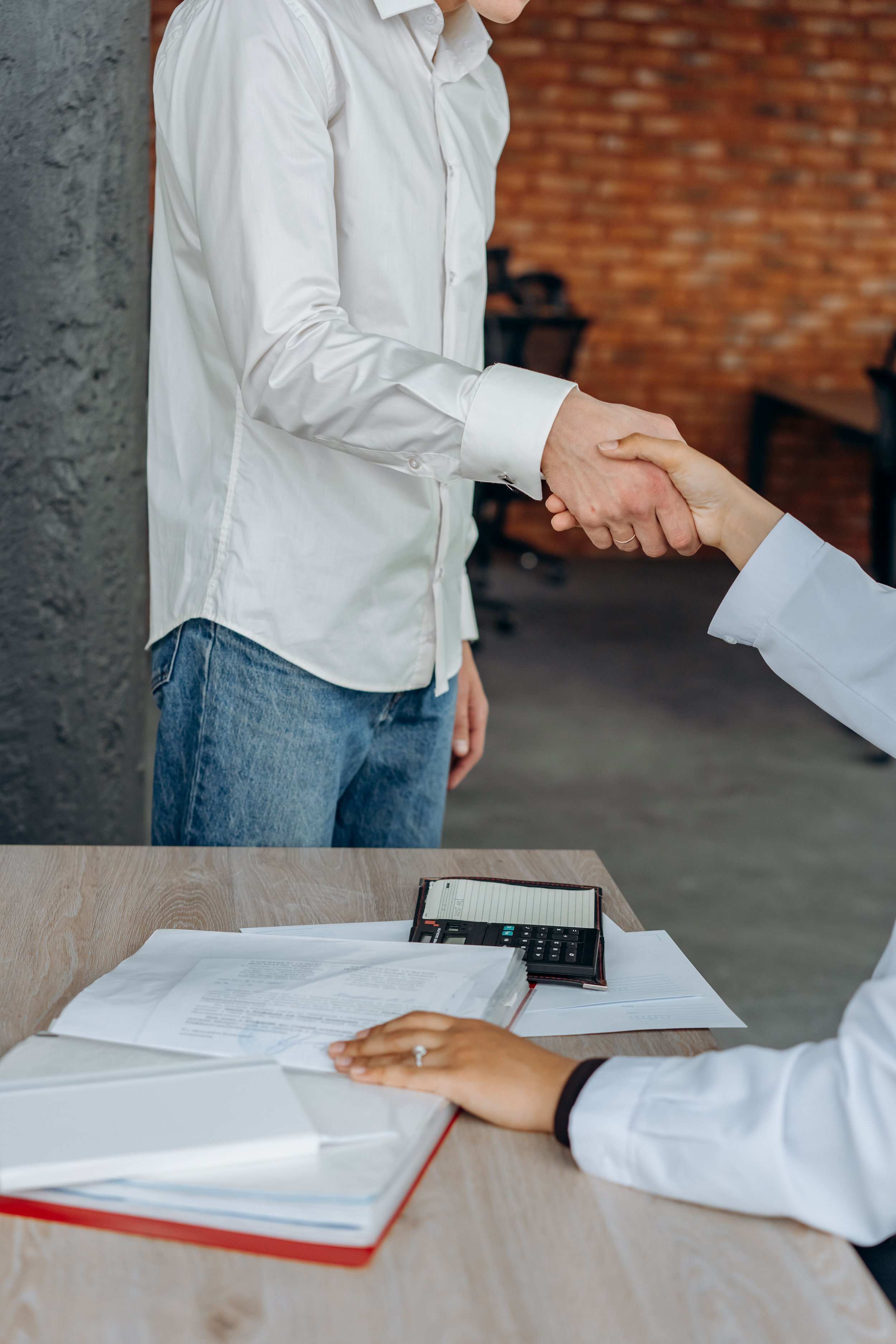 A person in a white shirt shaking hands with another person in a white blouse at a meeting or office setting. There are documents, a calendar, and a calculator on the table.