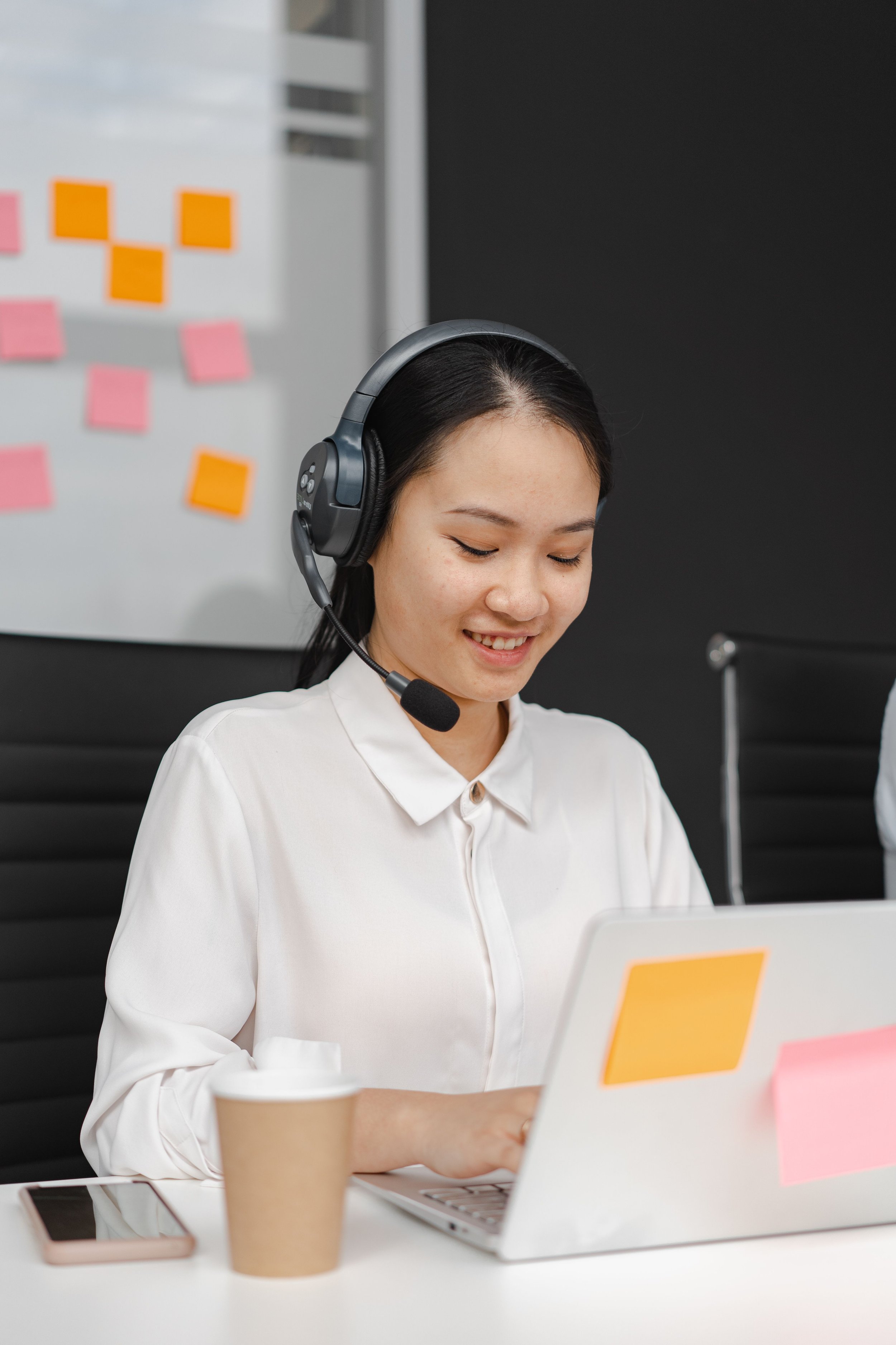 A woman with dark hair in a white shirt wearing a headset, smiling and looking at a laptop computer on a white desk with a coffee cup, a smartphone, and sticky notes in the background.