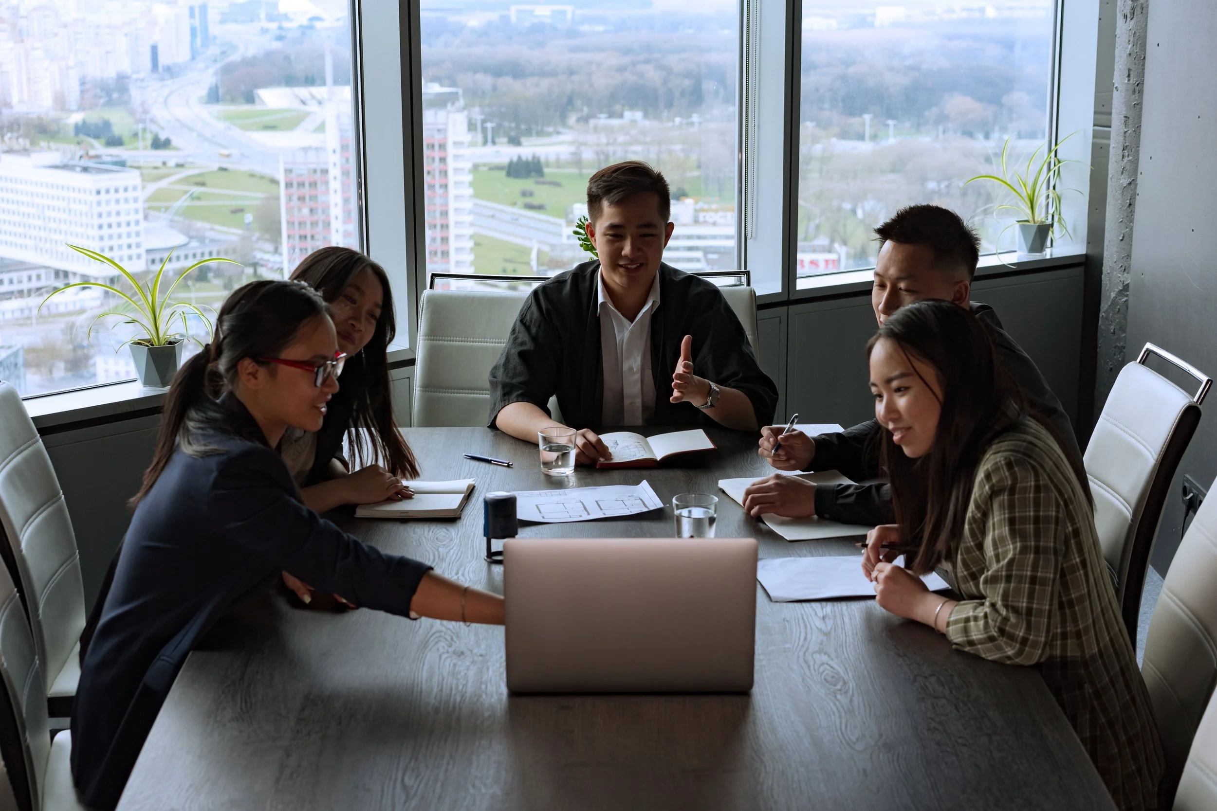 A group of six young professionals sitting around a conference table in a modern office with large windows showing a cityscape and skyline view. They are engaged in a discussion, with notebooks, pens, and a laptop on the table.